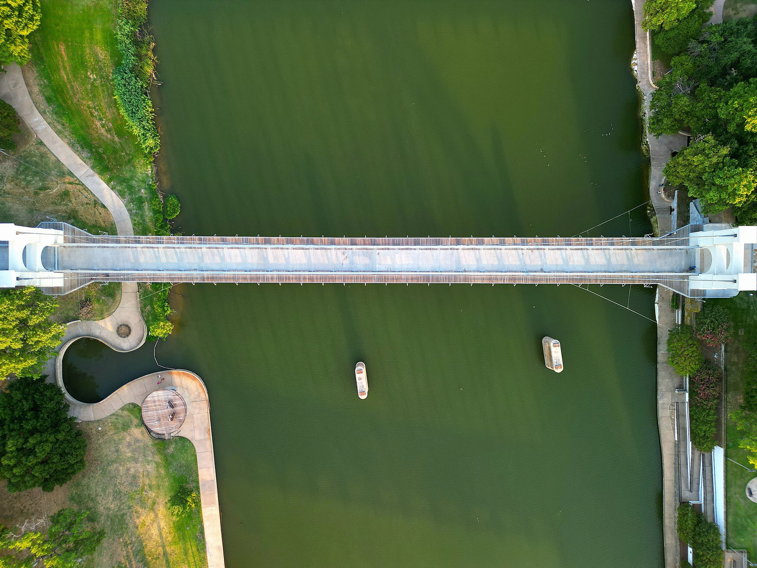 An aerial view of a suspension bridge crossing over a green body of water, with pathways, trees, and landscaped areas on either side.