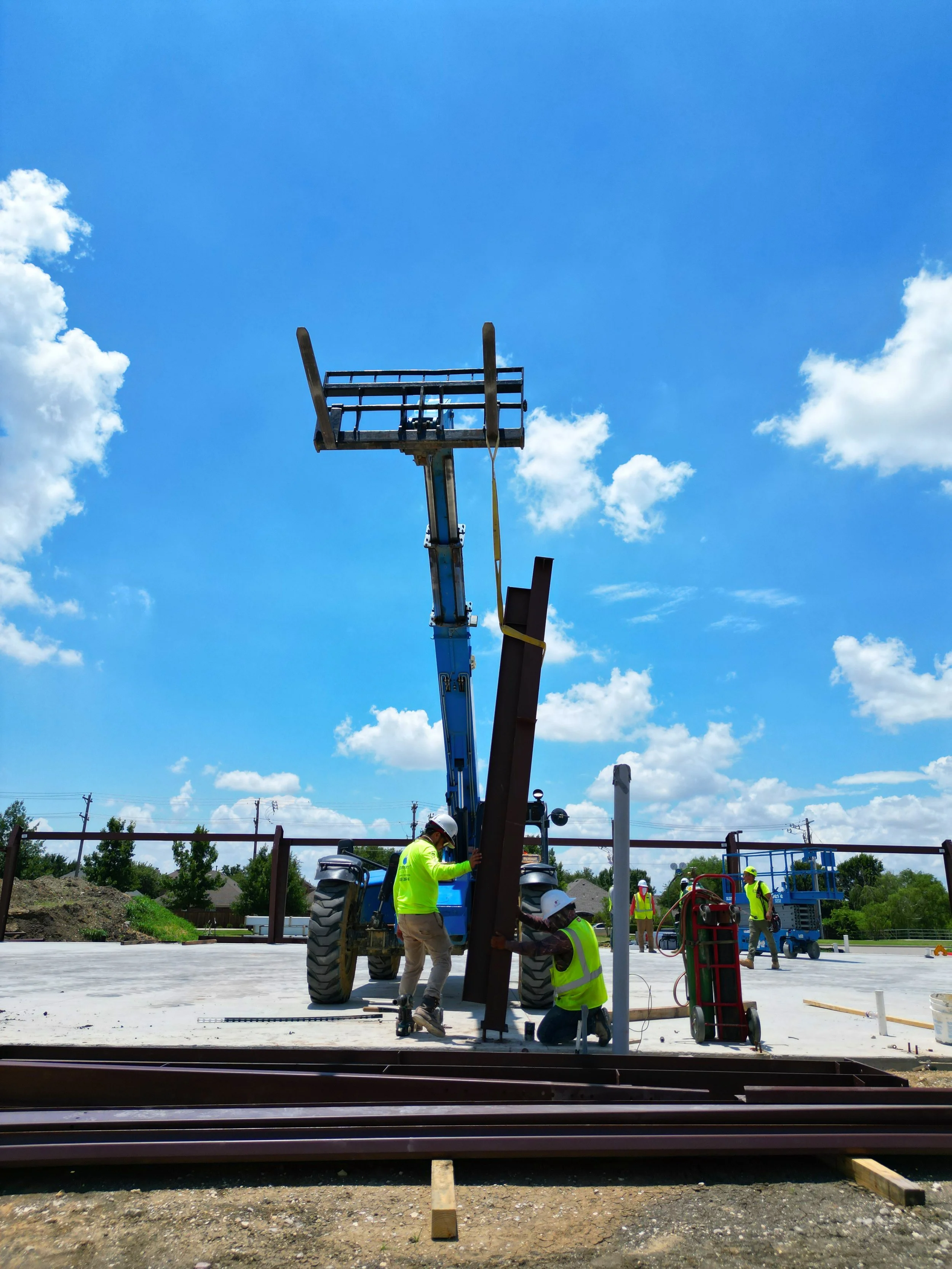 Construction workers in safety vests and helmets working on railroad tracks and steel beams at a construction site under a clear blue sky with scattered clouds.