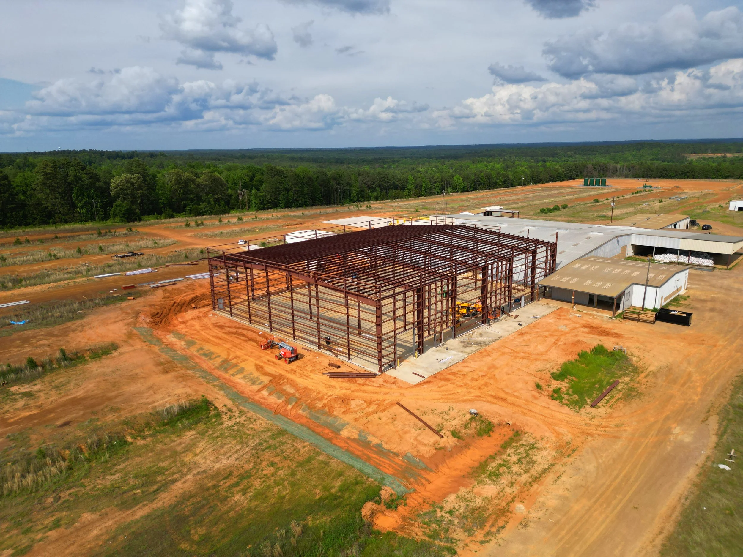 aerial view of a large construction site with a partially built steel frame structure surrounded by dirt, with trees and open land in the background and a partly cloudy sky overhead.