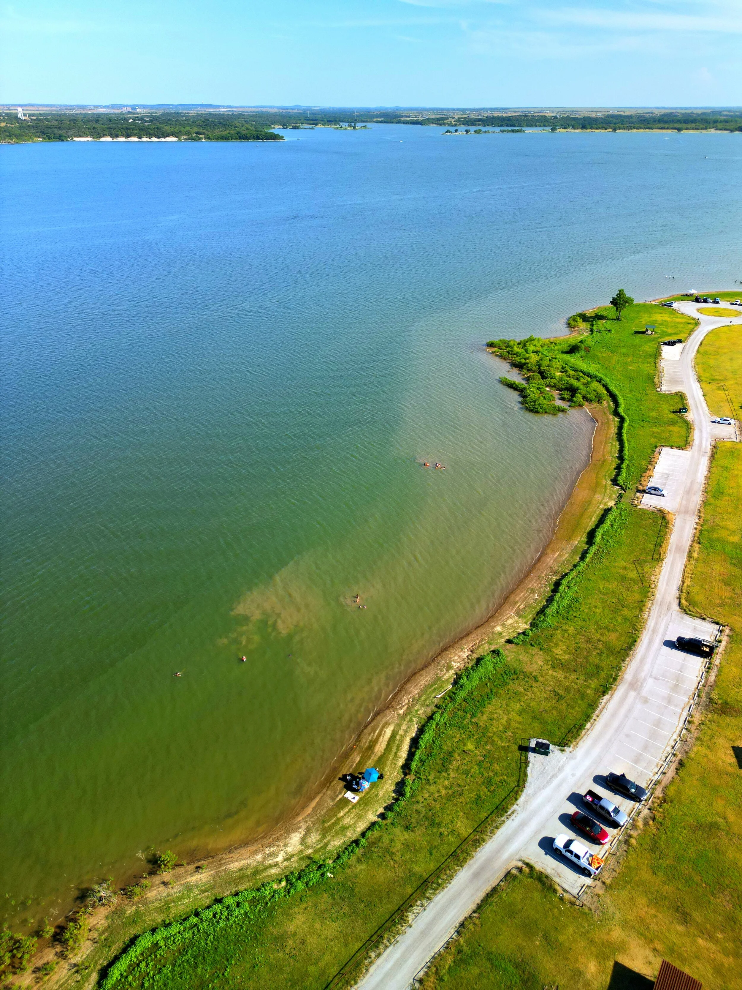 Aerial view of a large lake with clear blue water, a small shoreline area with some people swimming, and a parking lot with cars along a grassy area.