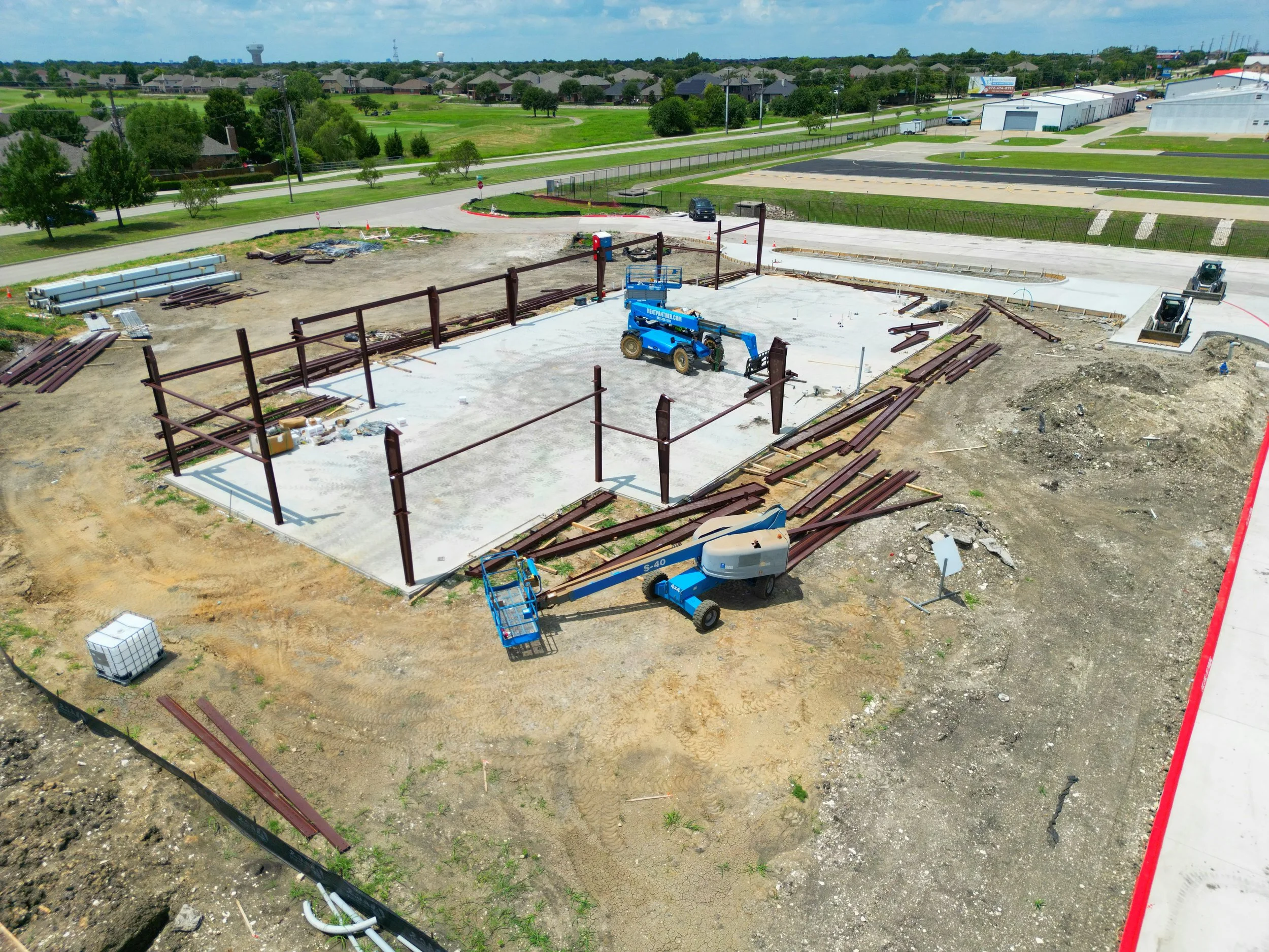Construction site with steel beams, a concrete foundation, a blue lift, and a cherry picker in a developing area with nearby roads and greenery.