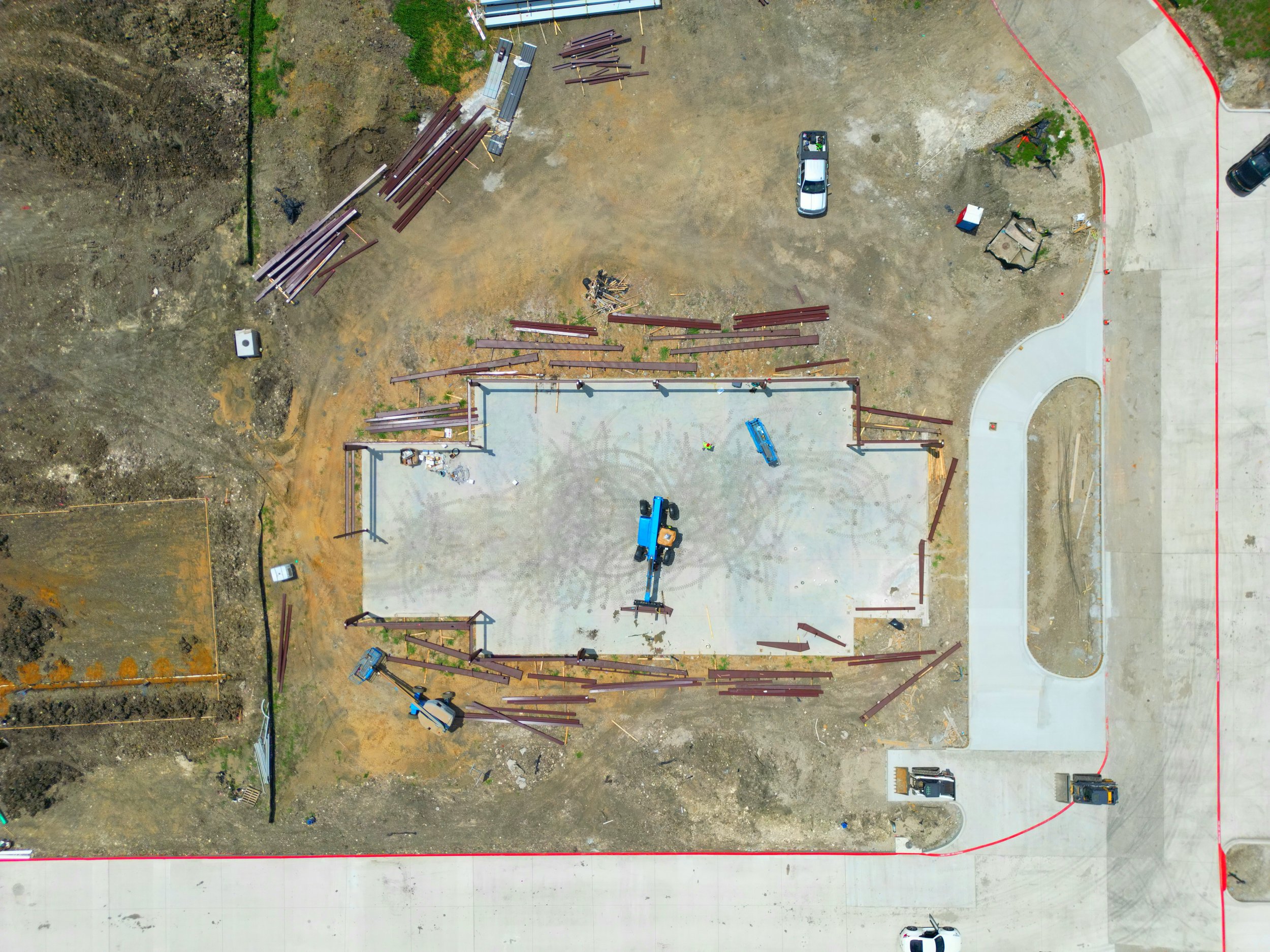 Construction site with a concrete foundation, steel beams, construction vehicles, and surrounding dirt and paved roads.