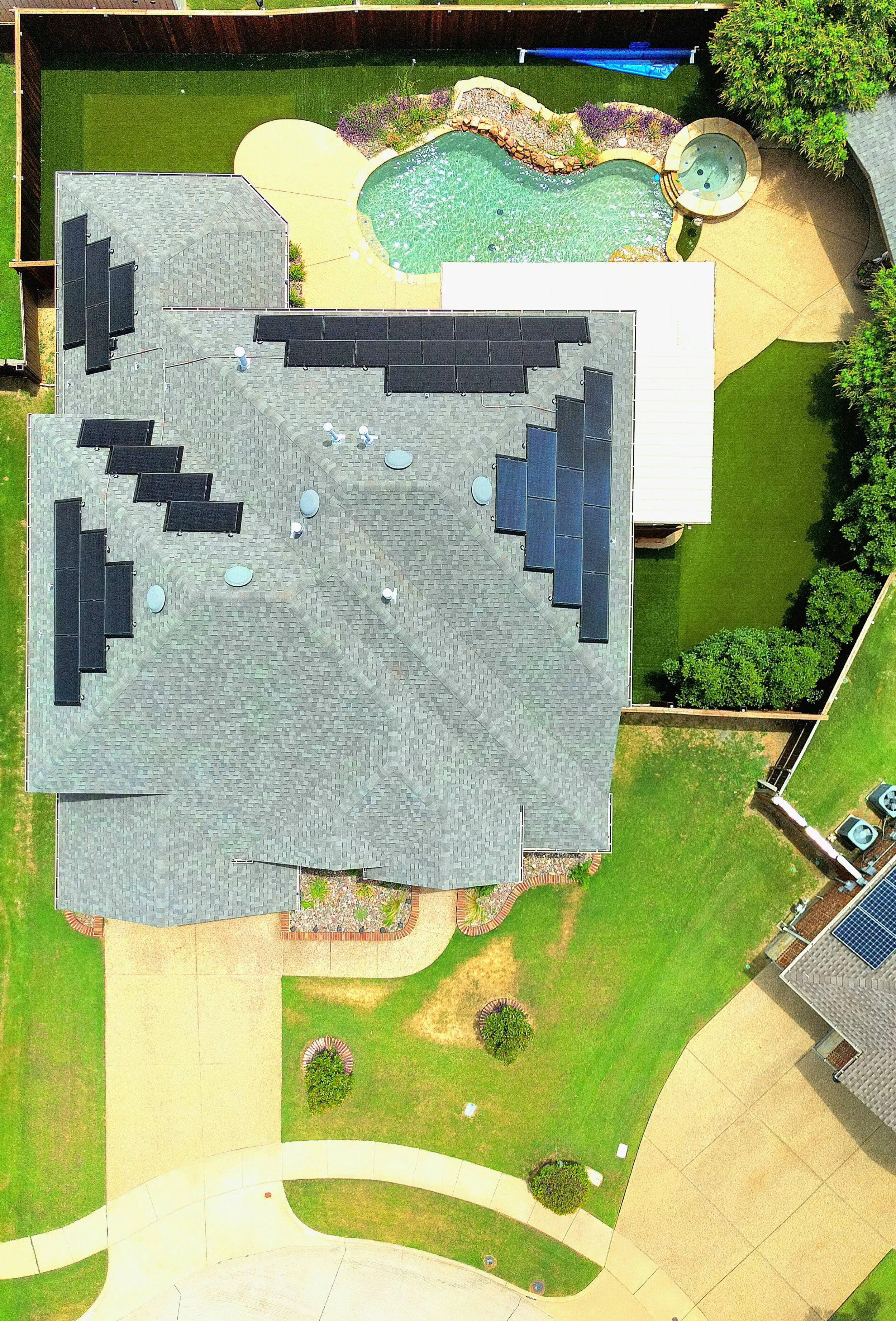 An aerial view of a house's backyard featuring a swimming pool with a small hot tub, a shaded patio area with solar panels on the roof, a manicured lawn with a curved sidewalk and a small landscaped garden, and neighboring houses with solar panels.