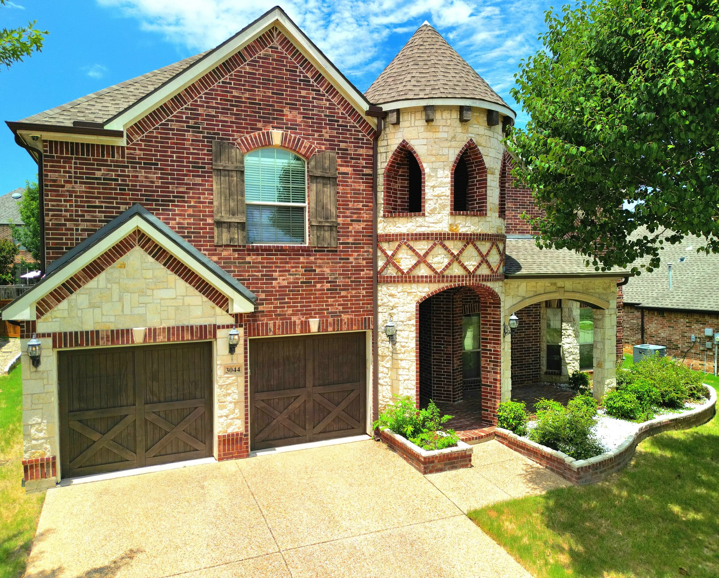 A two-story house with a brick and stone exterior, two garage doors, a small tower-like structure with arched windows, a front porch with a brick archway, and landscaped front yard with bushes and a sidewalk.