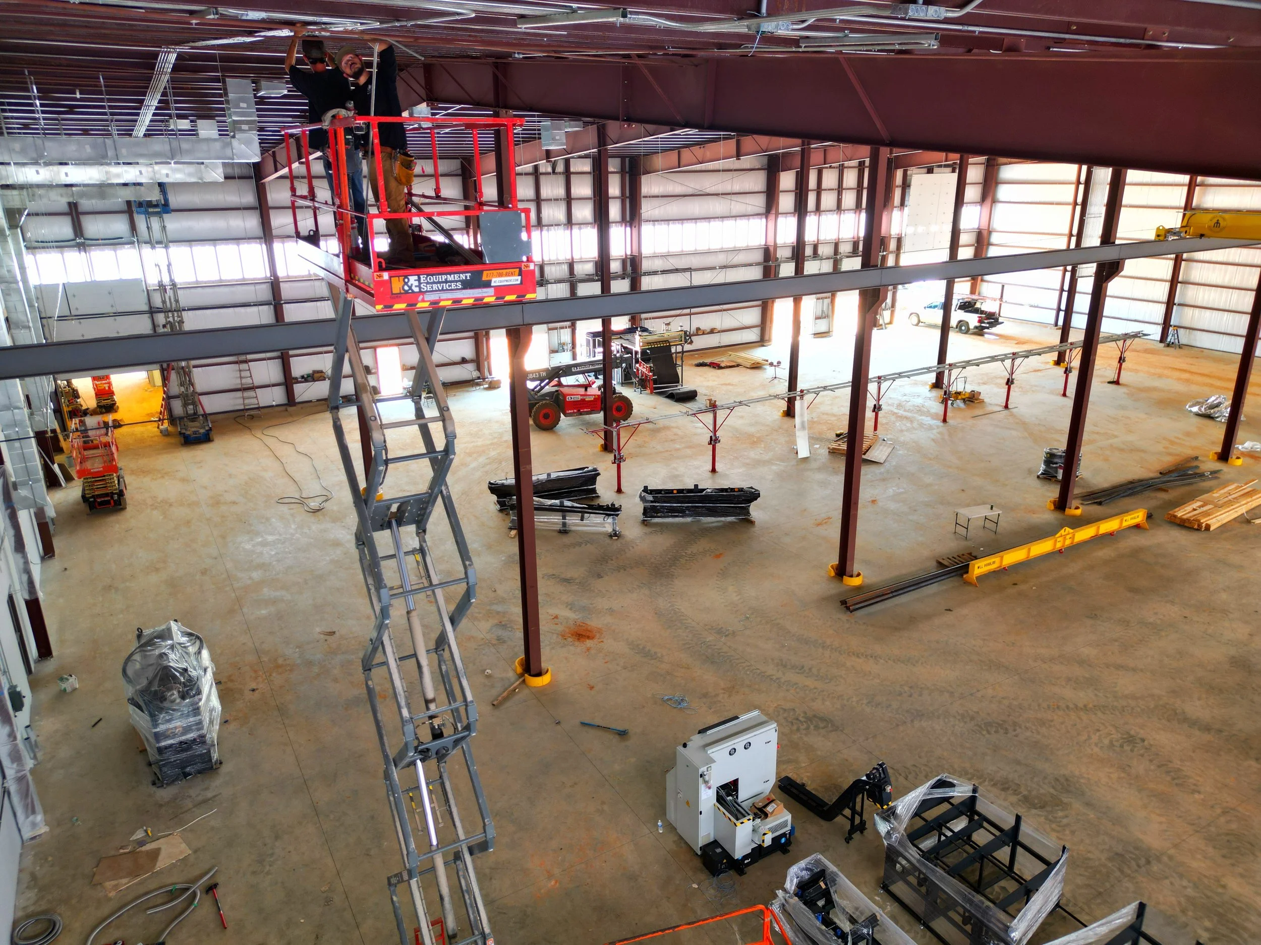 Interior view of a large construction warehouse with steel framework, construction equipment, and materials, with a worker in the elevated platform.