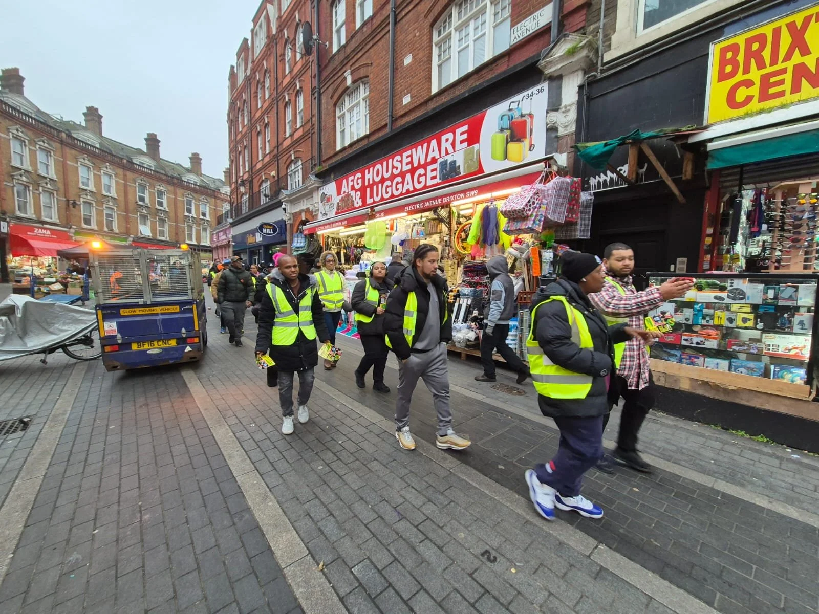 People wearing yellow reflective safety vests walking along a busy street with various shops, including a luggage and household store, in an urban area with brick buildings.