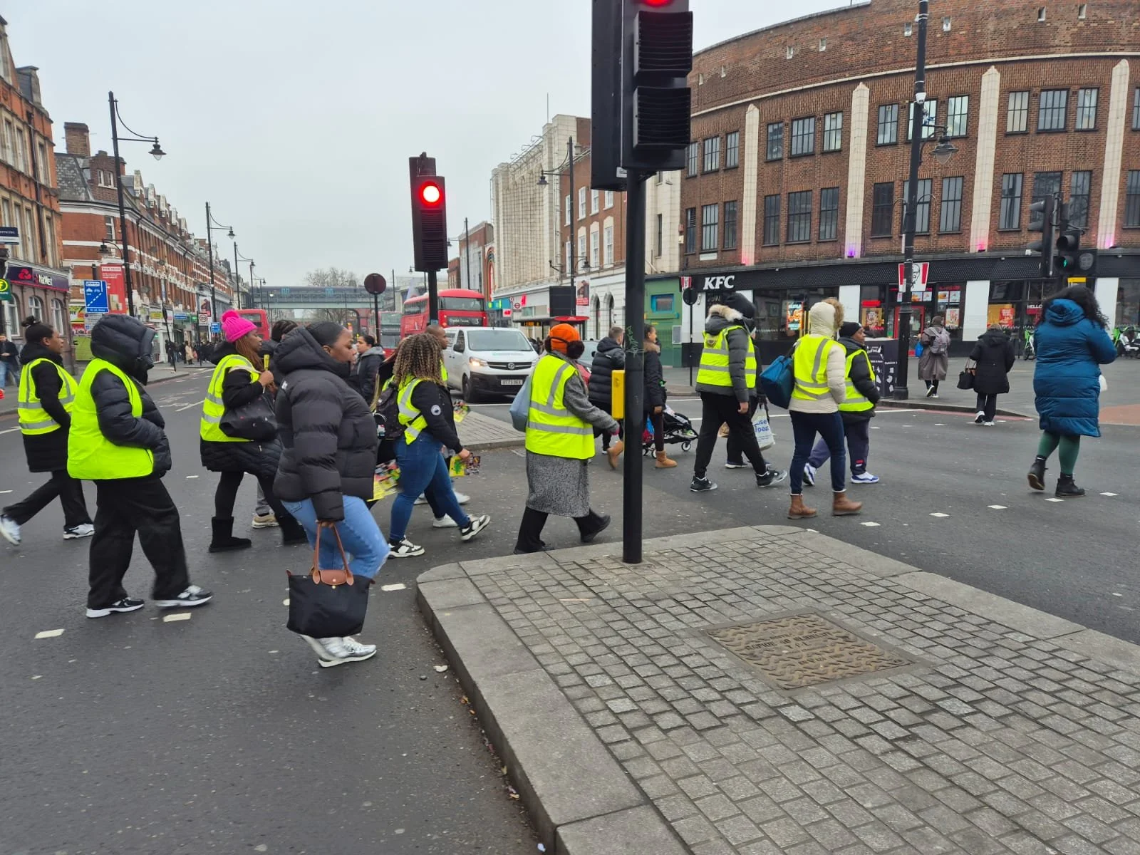 Crowd of people crossing a city street, some wearing yellow reflective vests, with traffic lights and shops in the background.