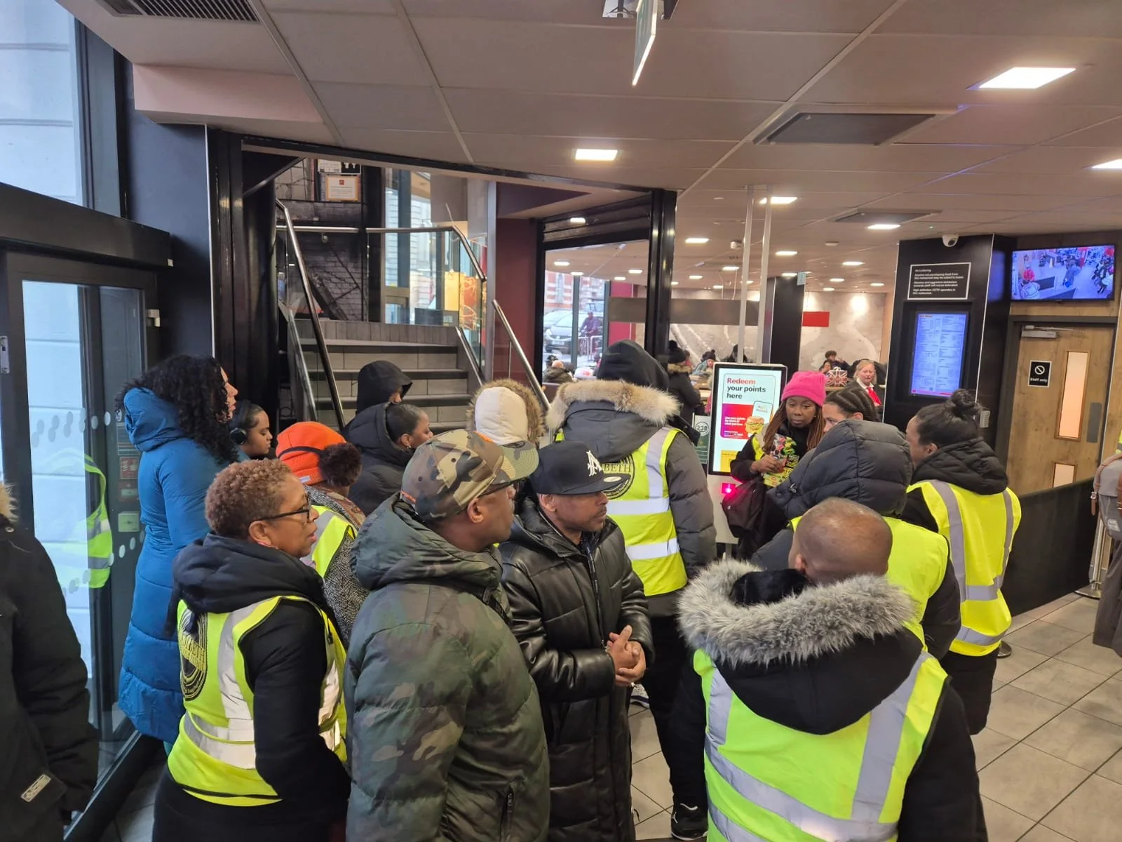 A group of people wearing yellow safety vests gathered inside a building, likely a restaurant or fast-food place, with some waiting in line and others talking to staff at a counter.