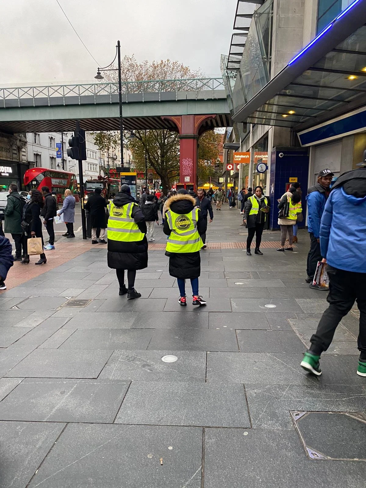A busy city street scene with pedestrians, some in high-visibility vests, and a red double-decker bus in the background. There is an overpass above, and a mix of modern and older buildings.