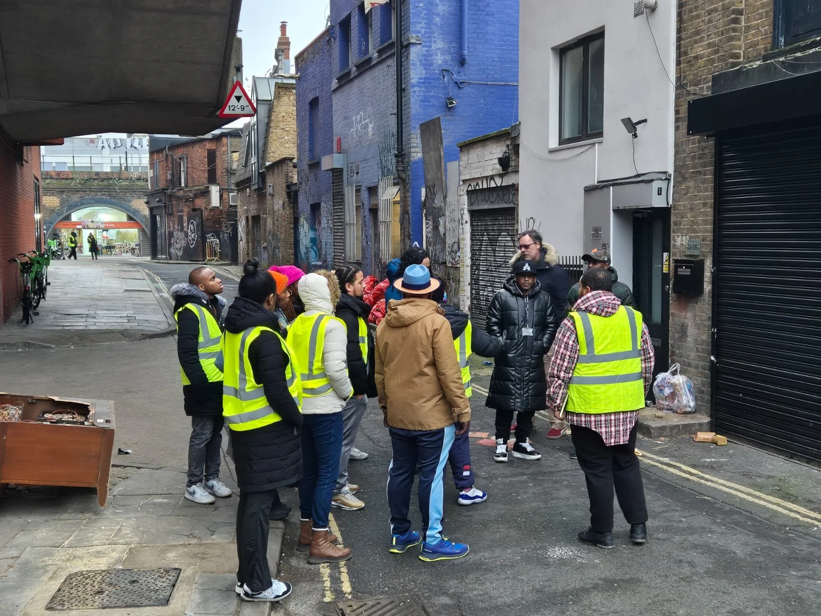 A group of people wearing yellow safety vests are gathered in an alleyway listening to a person speaking.