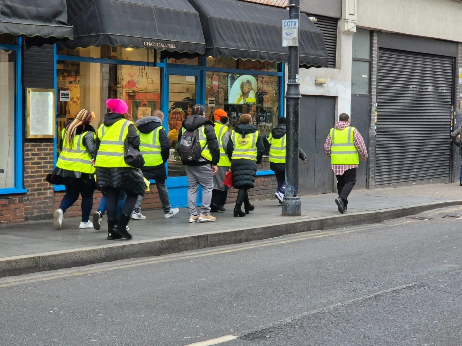 Group of people walking on sidewalk outside restaurant with bright yellow safety vests, some wearing backpacks and jackets, in an urban area.