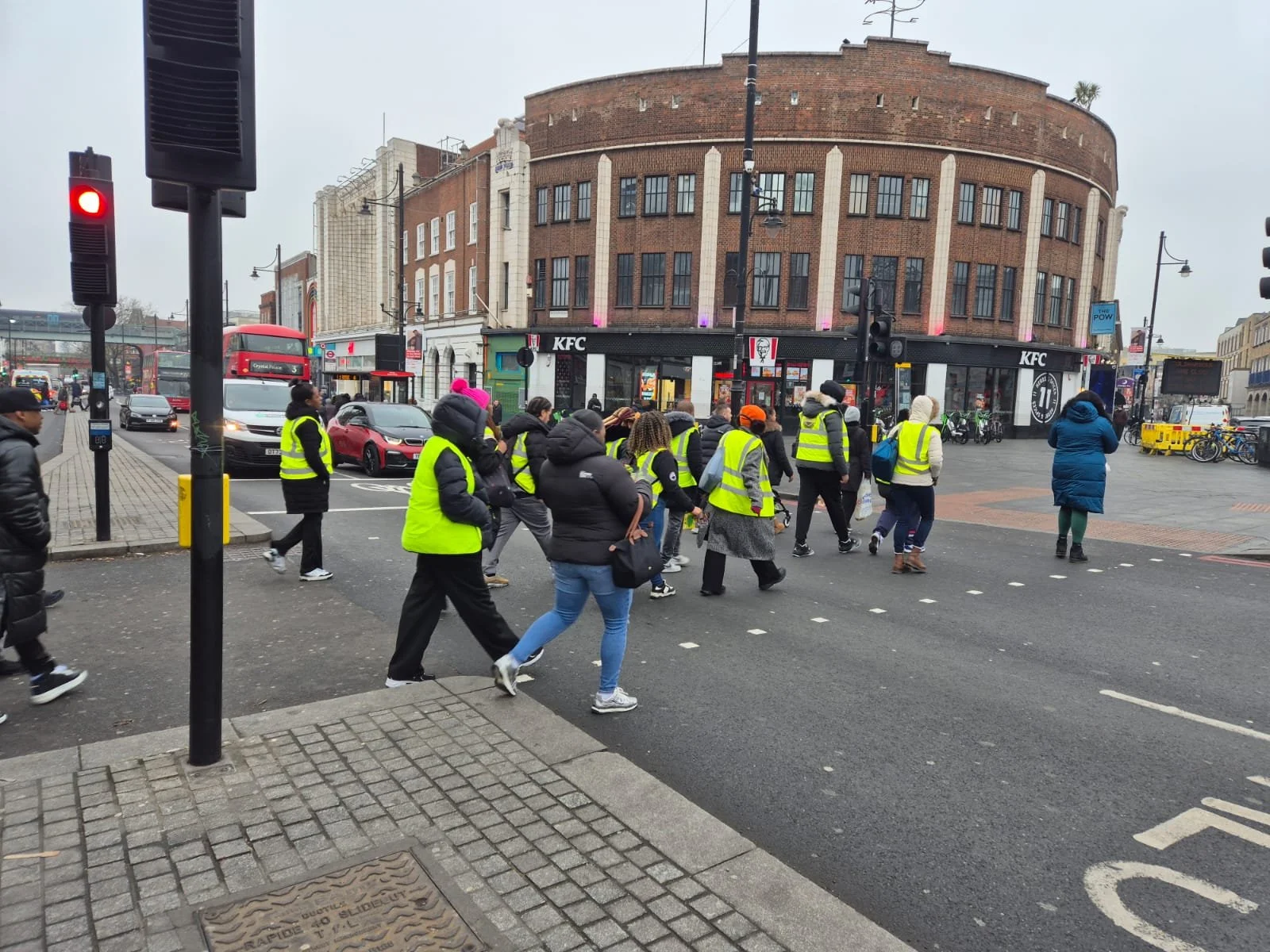 Crowd of pedestrians crossing the street at a busy intersection, some wearing yellow safety vests, in front of a KFC restaurant on a cloudy day.
