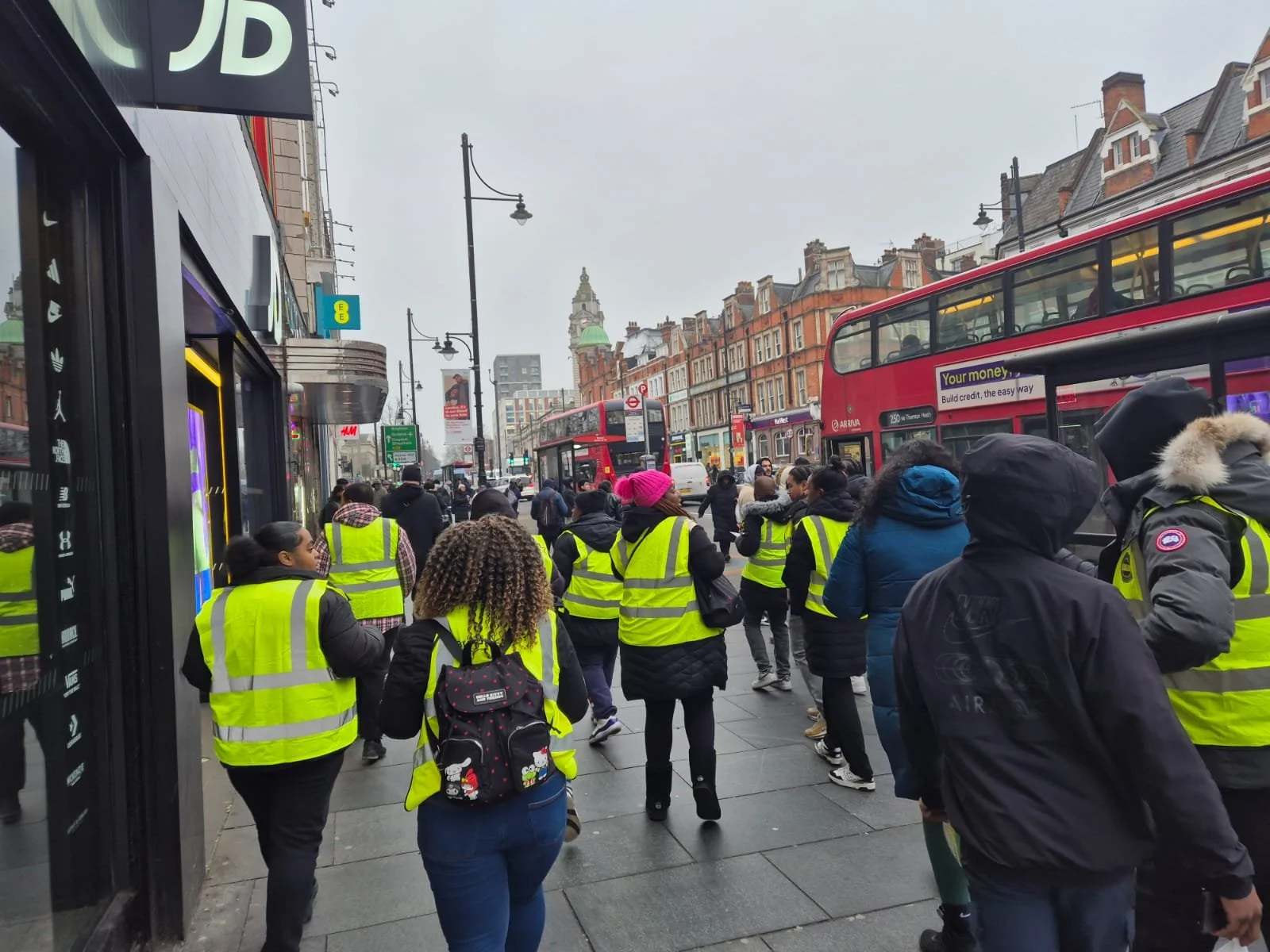 Crowd of people wearing yellow safety vests walking on a busy city sidewalk with red double-decker buses, buildings, and streetlights in the background.