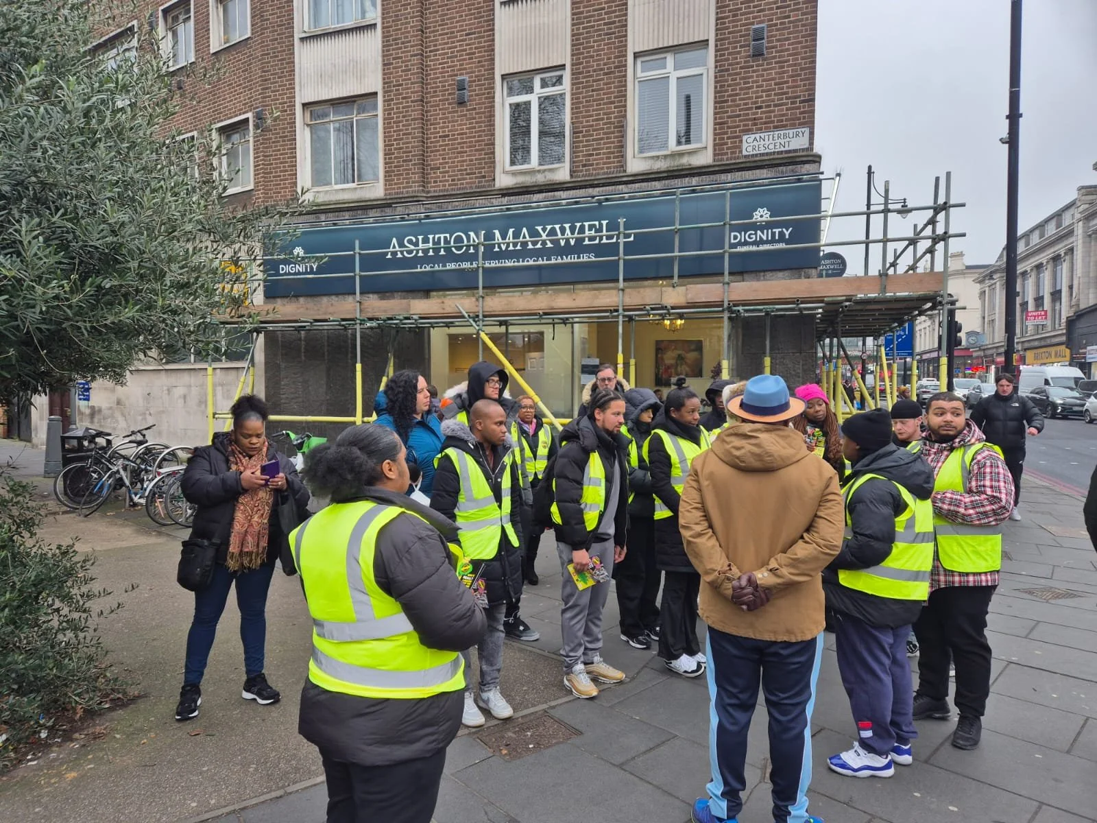 Group of people wearing yellow reflective vests gathered on city sidewalk in front of a building with scaffolding and a sign reading ASHTON MAXWELL, with bicycles parked nearby.