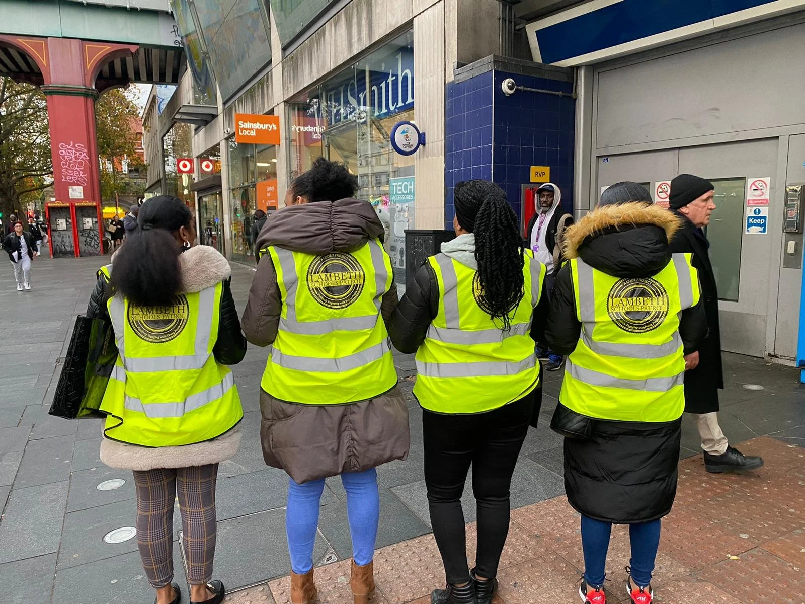 Group of five people wearing high-visibility jackets with 'Lambeth' logo, standing on a city sidewalk near storefronts and a street corner.