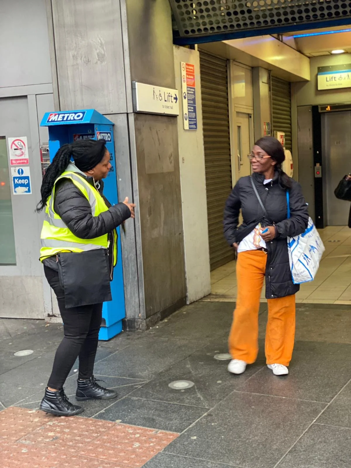 Two women talking outside a subway entrance. One woman wears a yellow safety vest, black jacket, black pants, and black boots, and is holding a black bag. The other woman wears an orange pair of pants, white shoes, glasses, and a black jacket, carryi