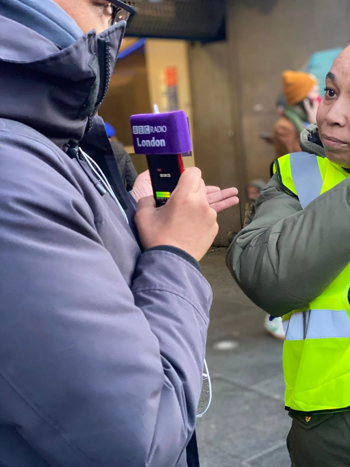 A person wearing a blue jacket holds a microphone labeled 'BBC Radio London' near another person wearing a high-visibility vest in an outdoor setting.