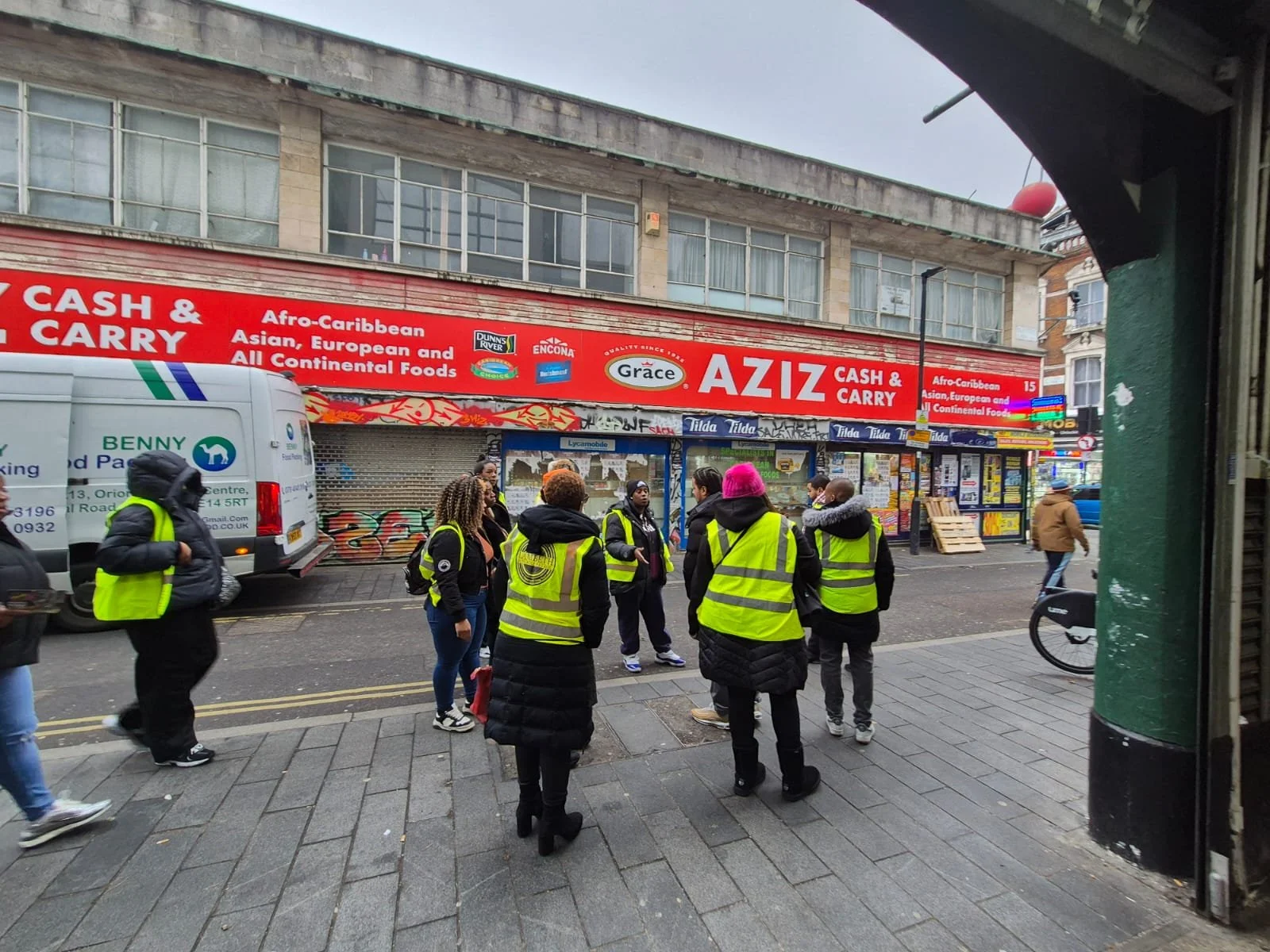 Group of people wearing yellow safety vests standing on a city sidewalk, with storefronts and a red sign reading "AZIZ CASH & CARRY" in the background.