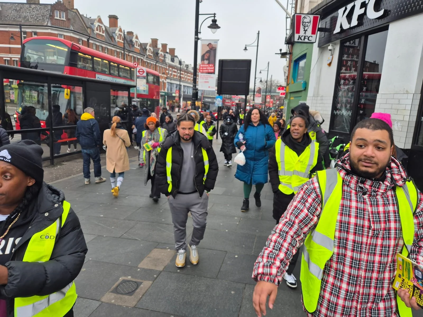 Crowd of people walking on city sidewalk, some wearing yellow safety vests, near a KFC restaurant and a red double-decker bus on a cloudy day.