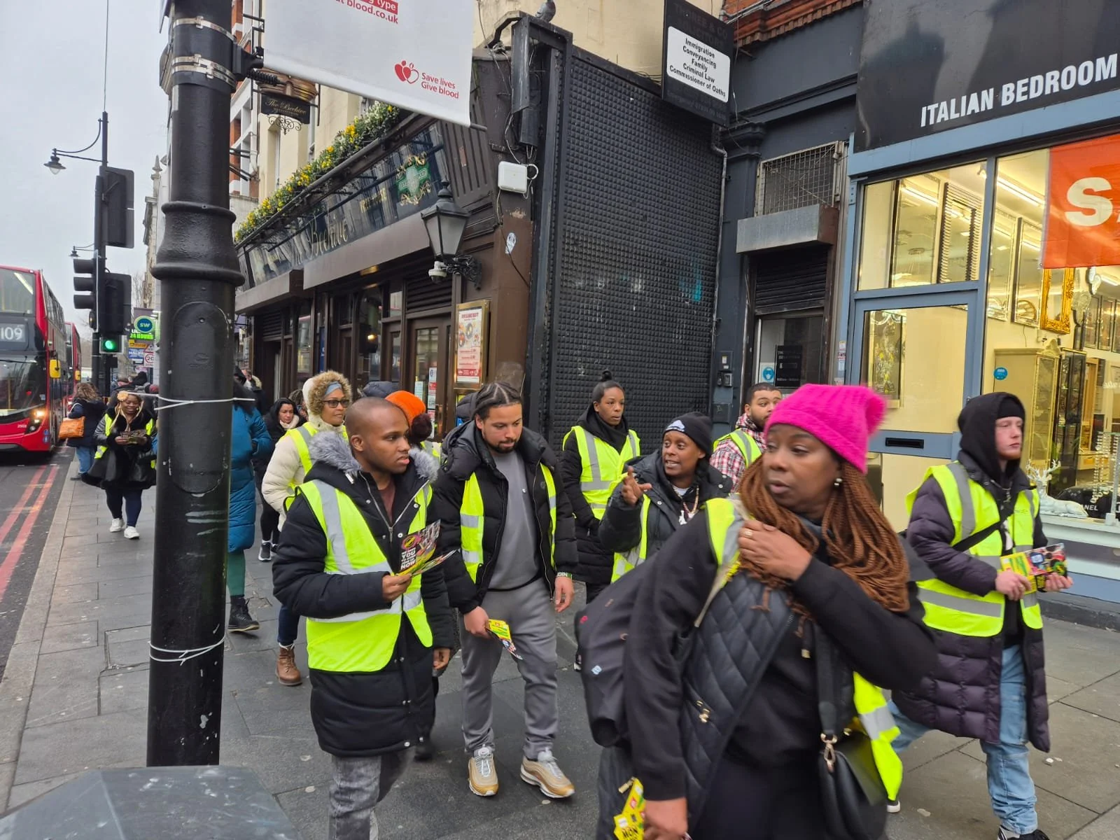 A group of people walking on a city sidewalk, many wearing yellow safety vests, some carrying pamphlets, with storefronts and a red bus in the background.