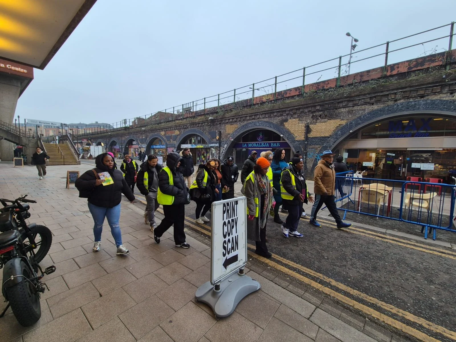 Group of people wearing yellow safety vests walking on a sidewalk in an urban area near shops and a railway underpass, with a sign that says 'PRINT COPY SCAN' in front of them.