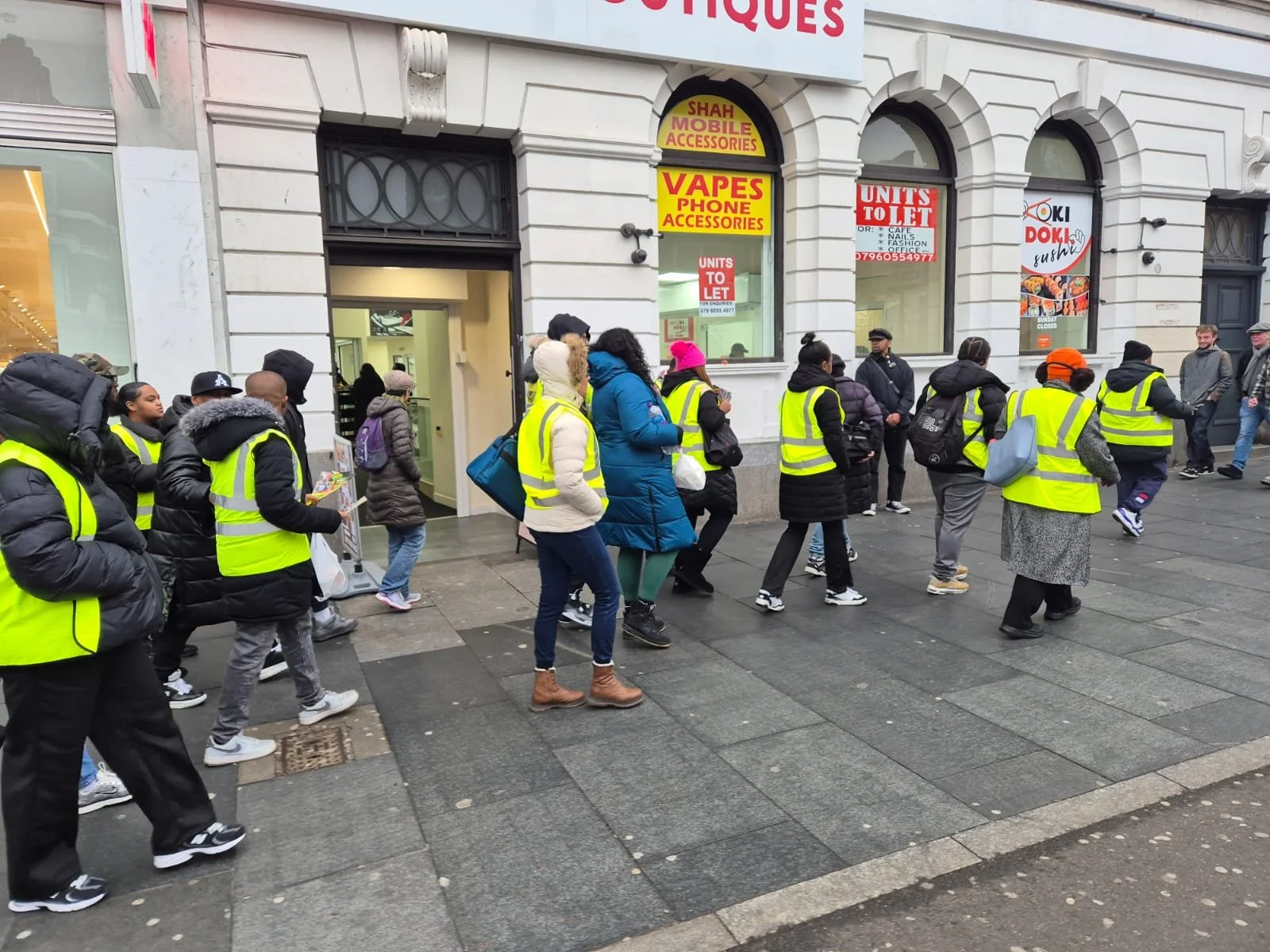 A line of people wearing yellow safety vests standing outside a shop on a city sidewalk.