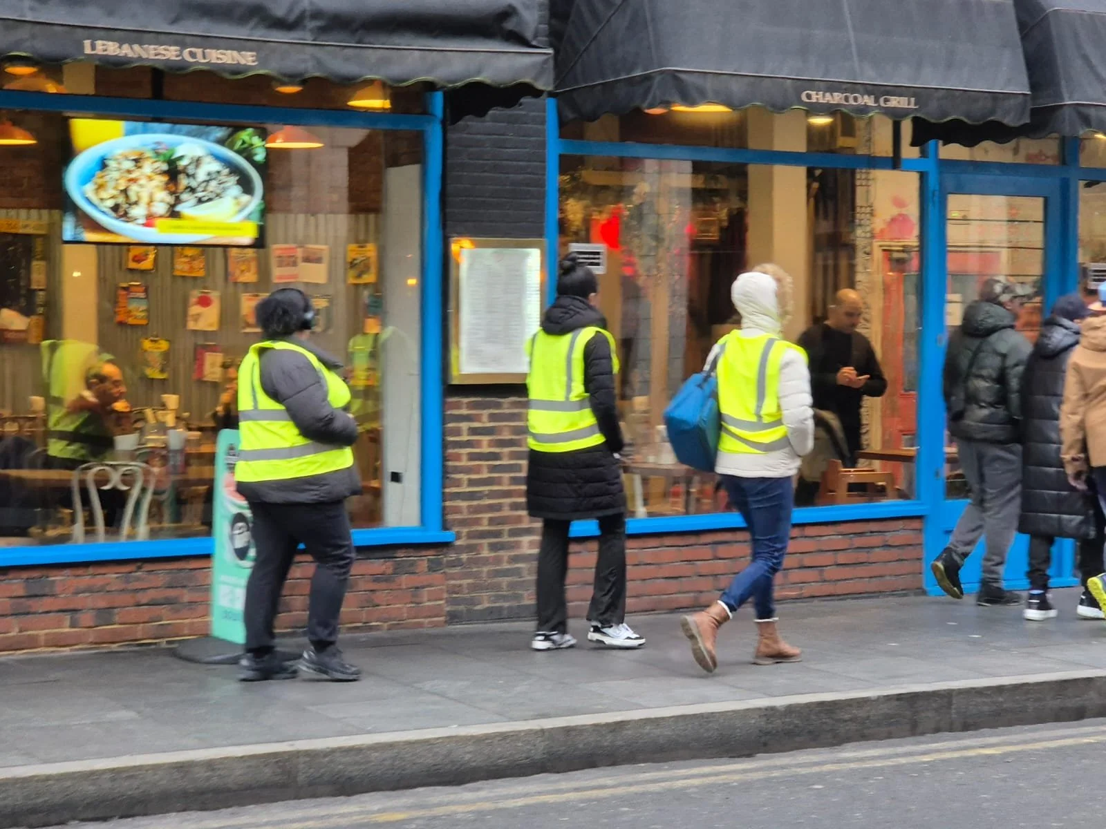 People wearing yellow safety vests standing in line outside a restaurant with brick and blue window frames, inside visible through glass windows.
