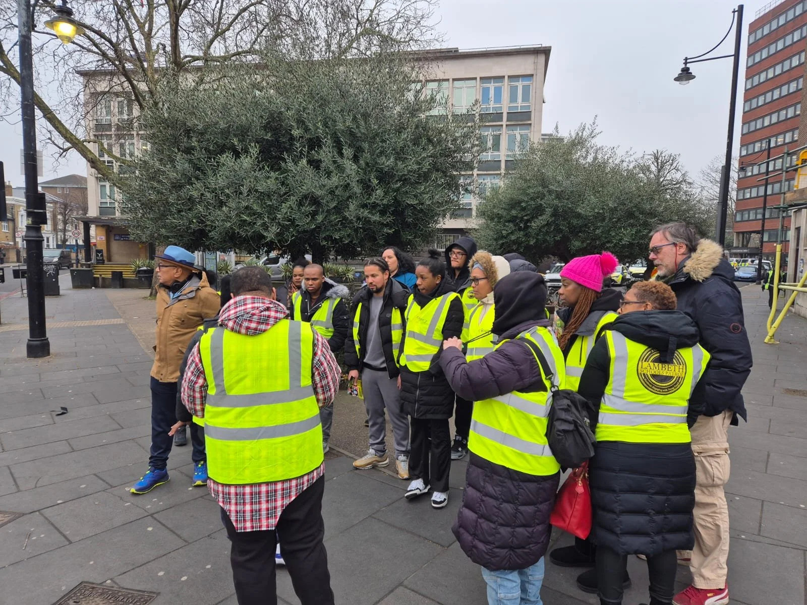 A group of people wearing yellow safety vests gathered outdoors on a city sidewalk, receiving instructions from a guide, with trees and buildings in the background.