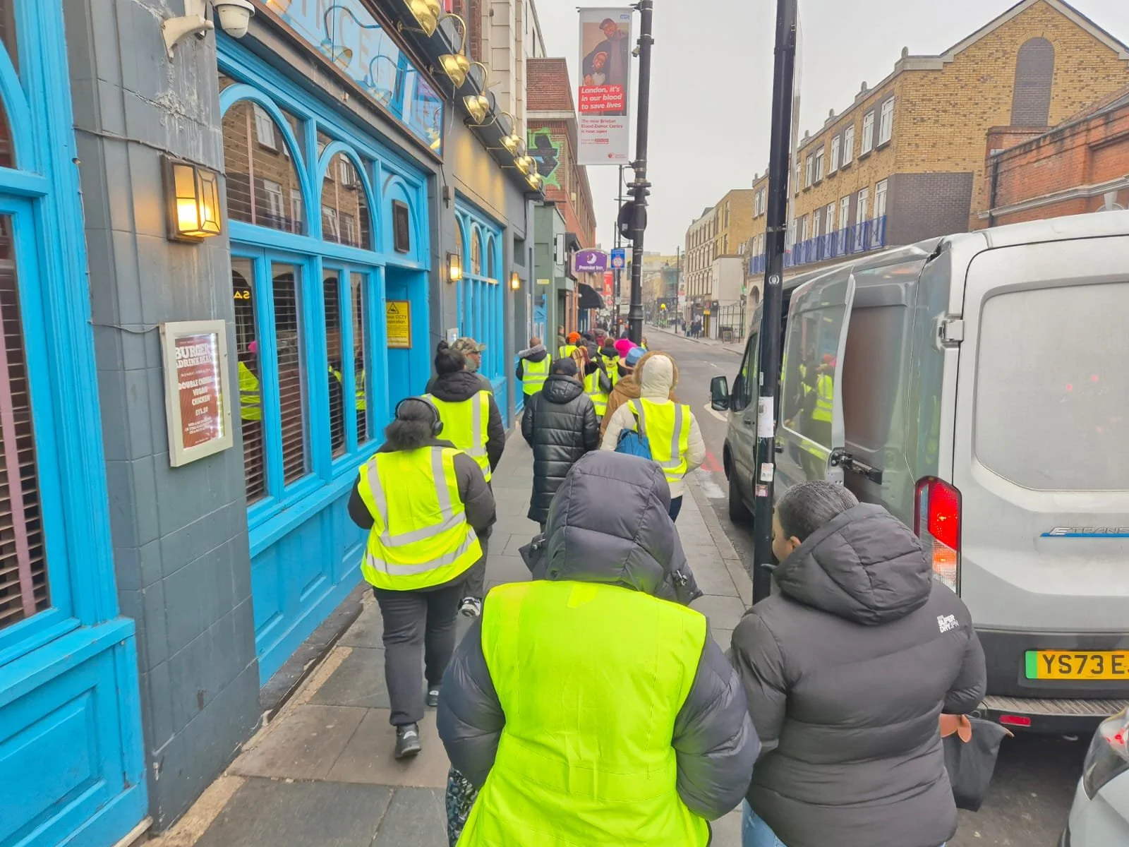A group of people wearing yellow safety vests walking in a line on a city sidewalk next to blue buildings and parked vehicles.