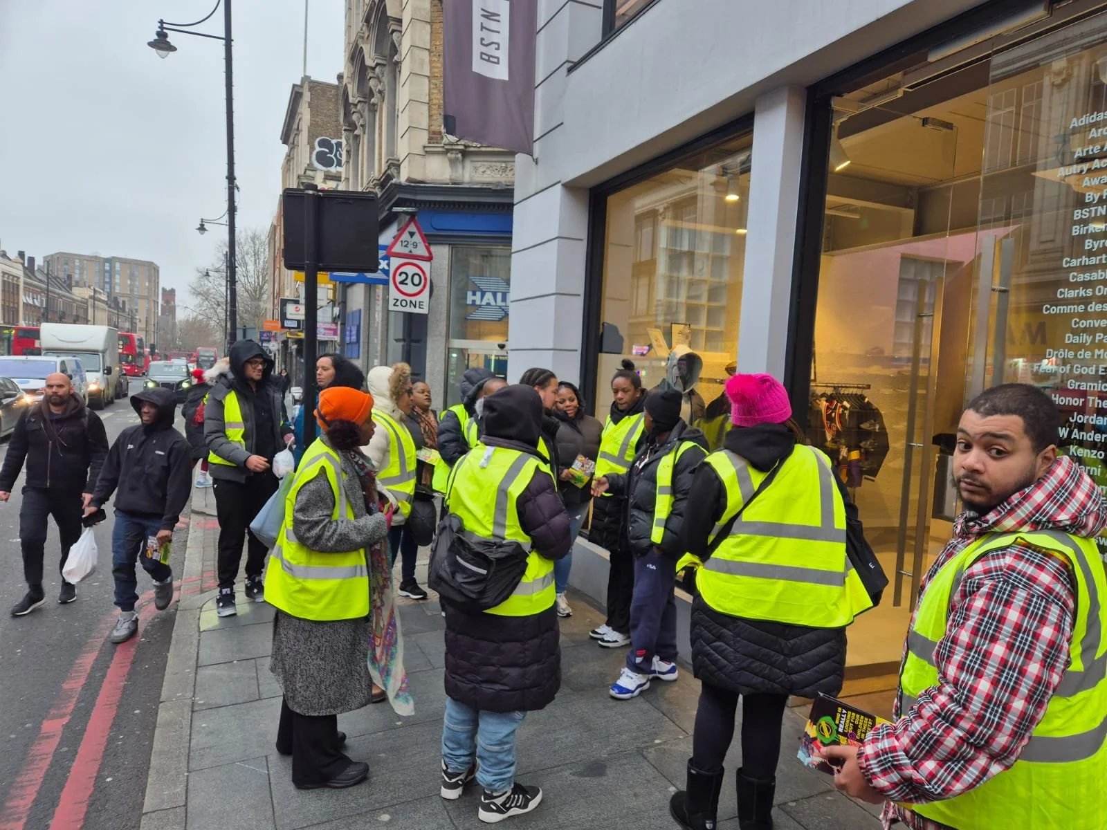 A group of people wearing yellow safety vests standing on the sidewalk outside a store, waiting in line or gathering near the entrance.