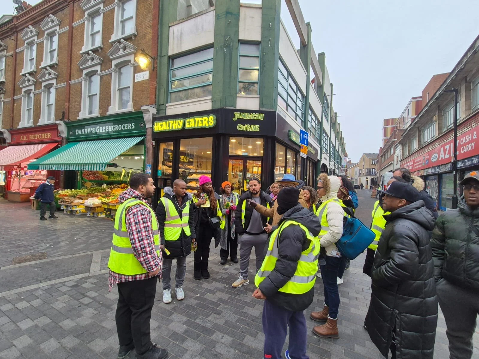 A group of people wearing yellow safety vests gathered on a city street corner, with storefronts including a grocery store with fruit displays and a restaurant named 'Healthy Eaters' in the background.