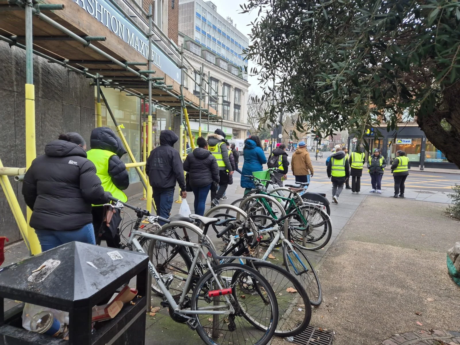 People, many wearing yellow safety vests, waiting in line outside a building under construction with bicycles parked nearby in an urban setting.