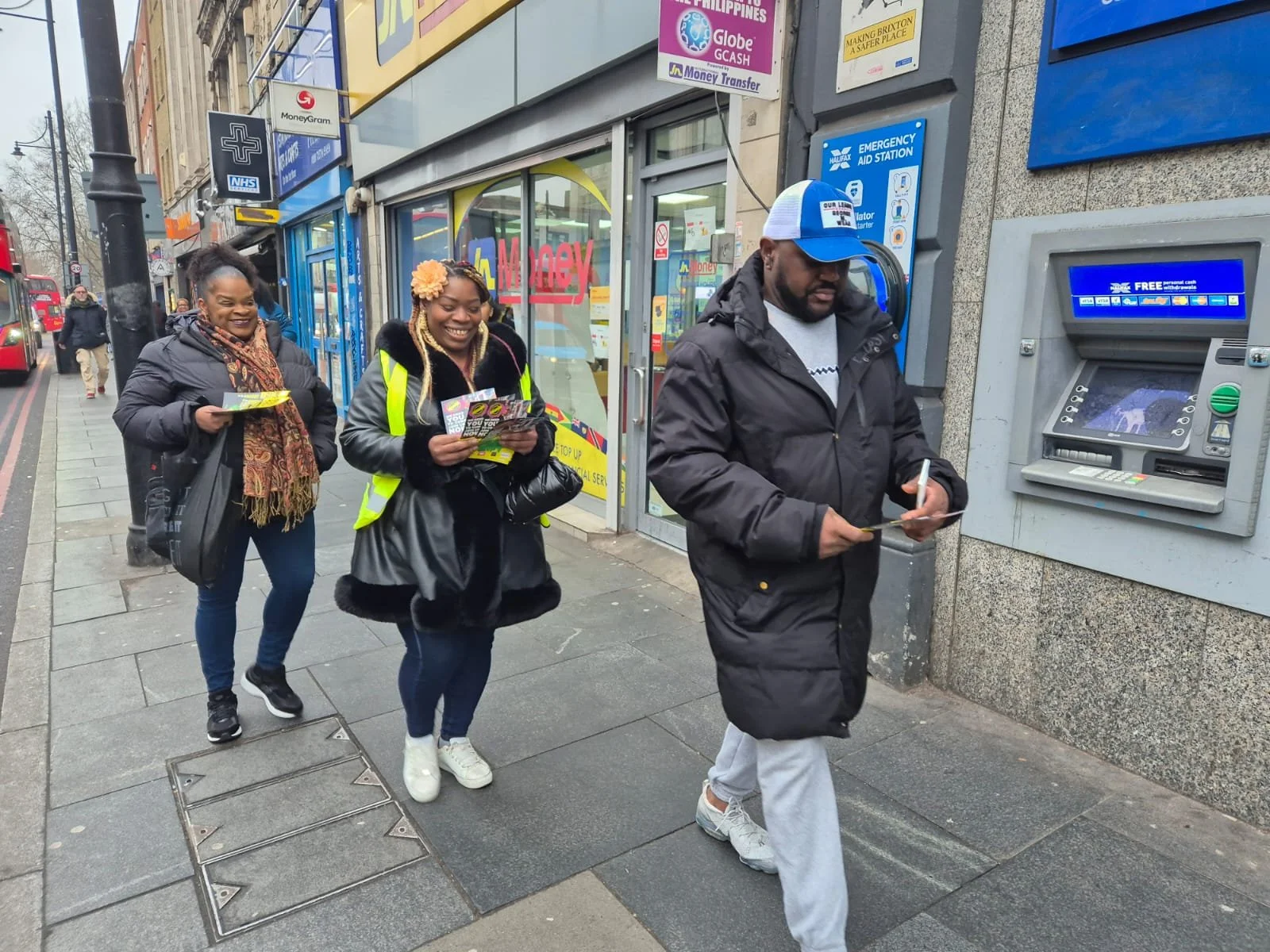 Three women and one man walking on a city sidewalk, with the man using an ATM machine. The women are smiling and holding flyers or brochures, and they are dressed in casual clothing with jackets. The man is wearing a black jacket, gray sweatpants, an