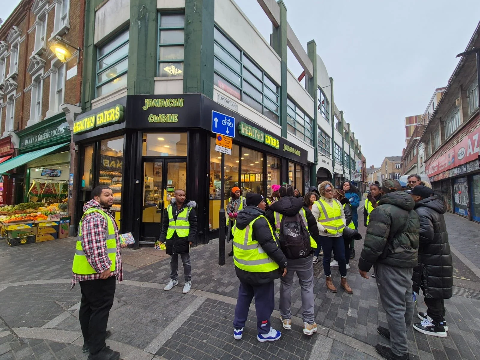 Group of people wearing yellow safety vests standing on a city sidewalk outside a Jamaican cuisine restaurant called Healthy Eaters, with fruit stalls nearby.