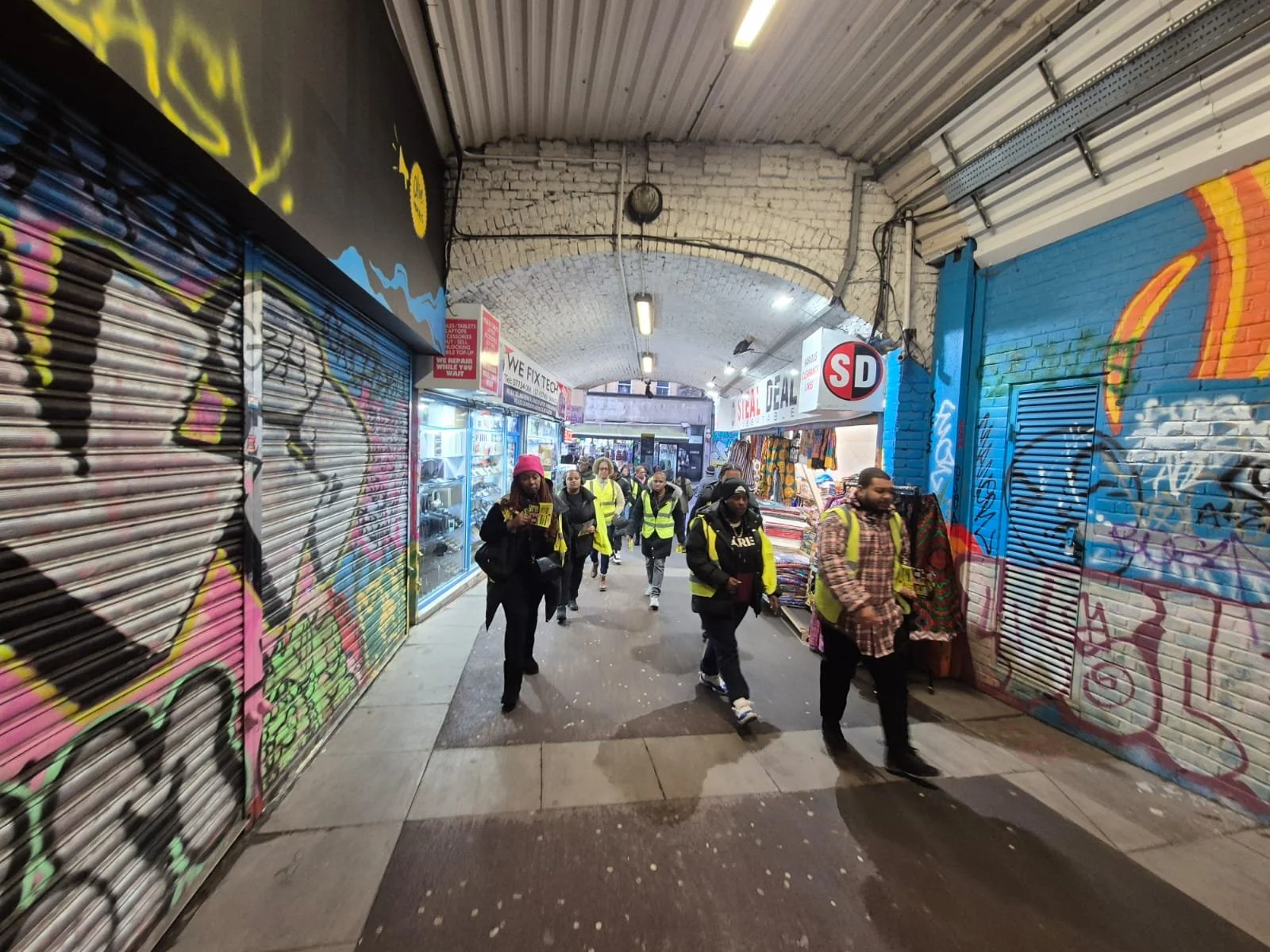 People walking through a covered marketplace with graffiti-covered metal shutters on the sides, some of them wearing yellow safety vests. The marketplace has brick arches and a variety of shops visible in the background.