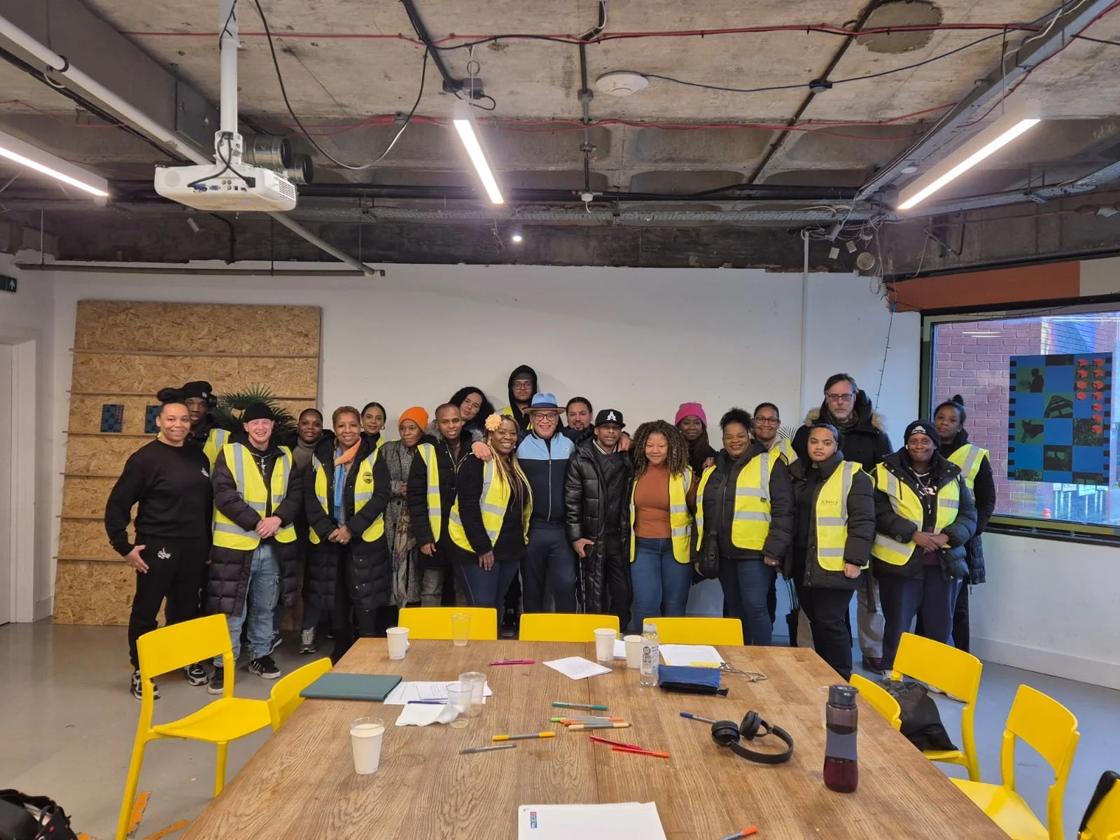 A group of approximately 20 people wearing yellow safety vests posing indoors in front of a white wall with a large wooden panel, in a room with industrial ceiling and fluorescent lighting, next to a window with colorful stained glass.