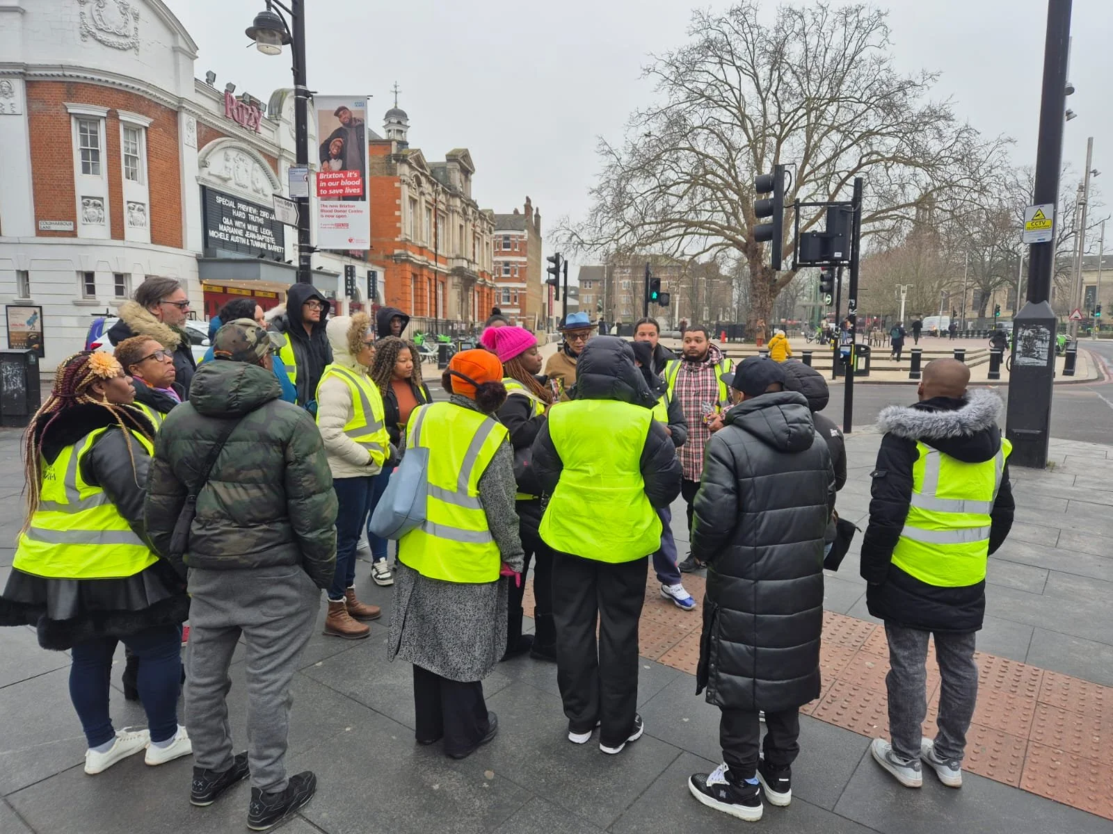 Group of people with yellow safety vests gathered for a discussion on a city street corner, with buildings, traffic lights, and leafless trees in the background.