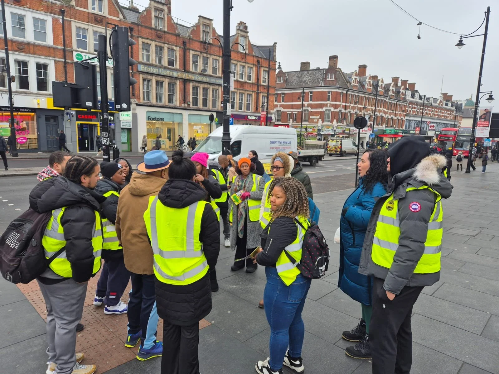 A group of people, mostly women, wearing yellow reflective vests, gathered on a city sidewalk for a discussion or tour on a busy street. There are shops and buildings in the background, as well as vehicles and pedestrians.