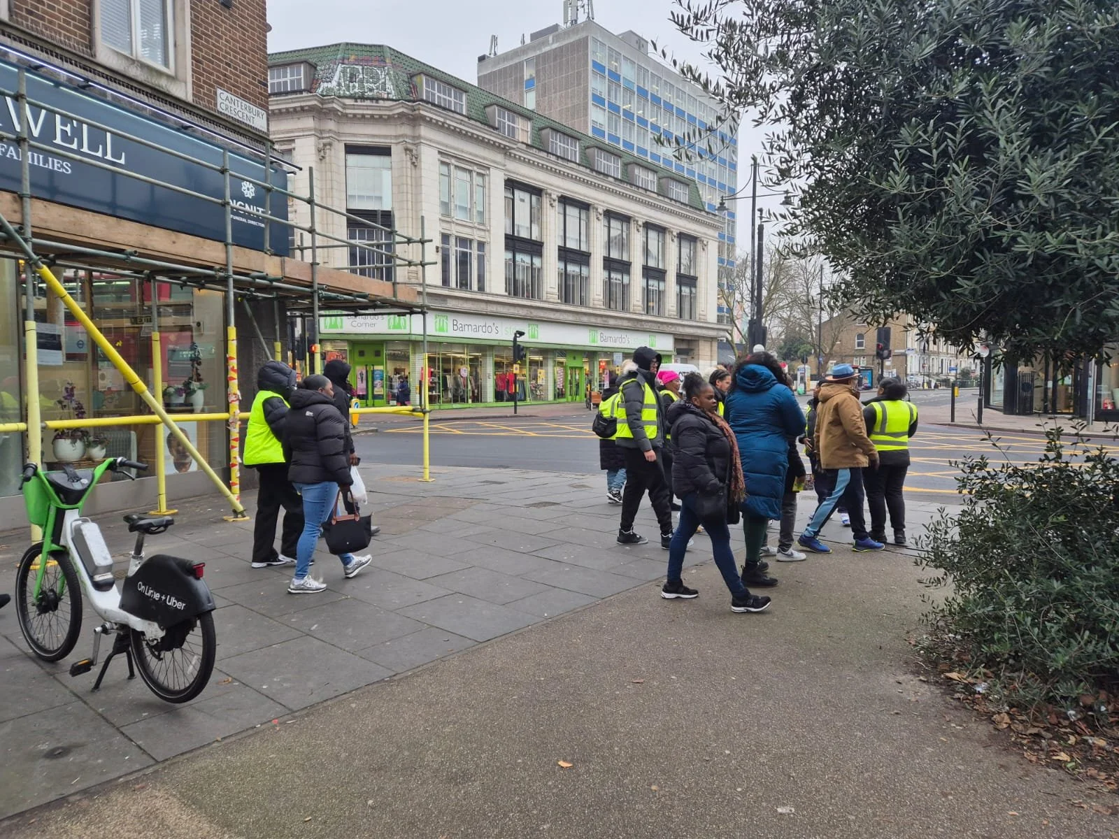 A group of people waiting to cross the street at a city intersection, some wearing reflective vests, with buildings and storefronts in the background.