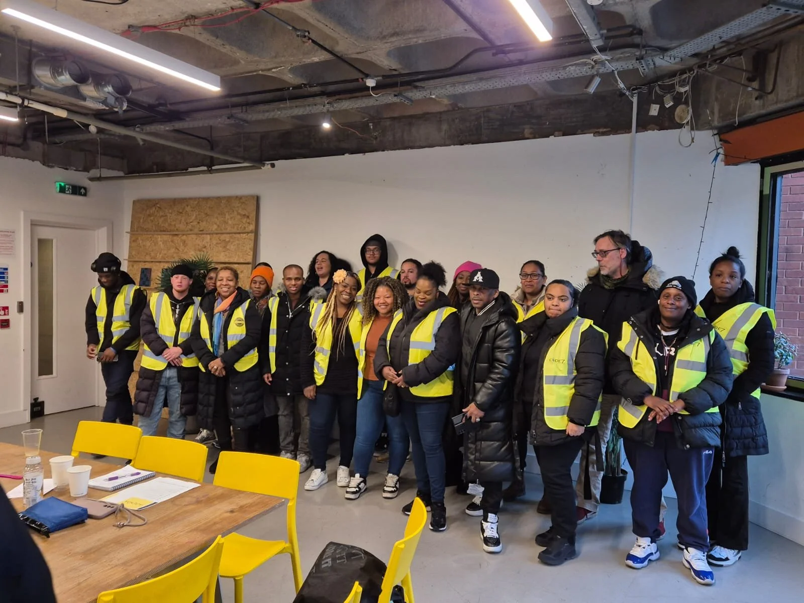 Group of people wearing yellow safety vests inside a room with a concrete ceiling, some sitting at yellow tables, smiling for the photo.