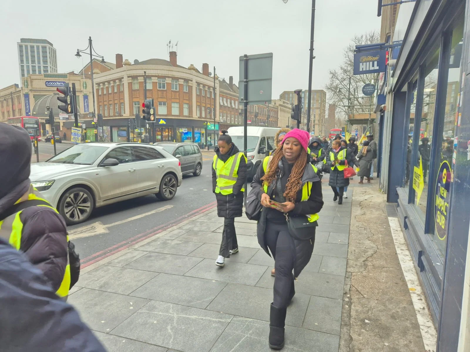 People walking on a city sidewalk with some wearing yellow reflective vests, near traffic lights and cars. The buildings in the background are multi-story with shops on the ground level.