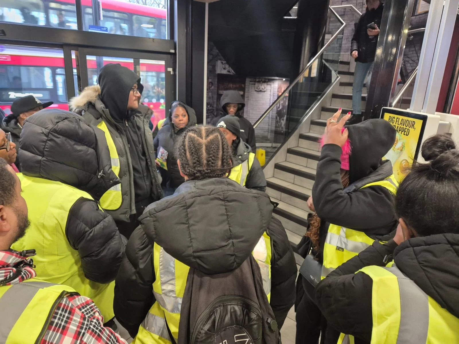 Group of people, mostly women, wearing yellow safety vests, gathered indoors near a staircase, some wearing hooded jackets, in a busy location with large windows and a staircase.