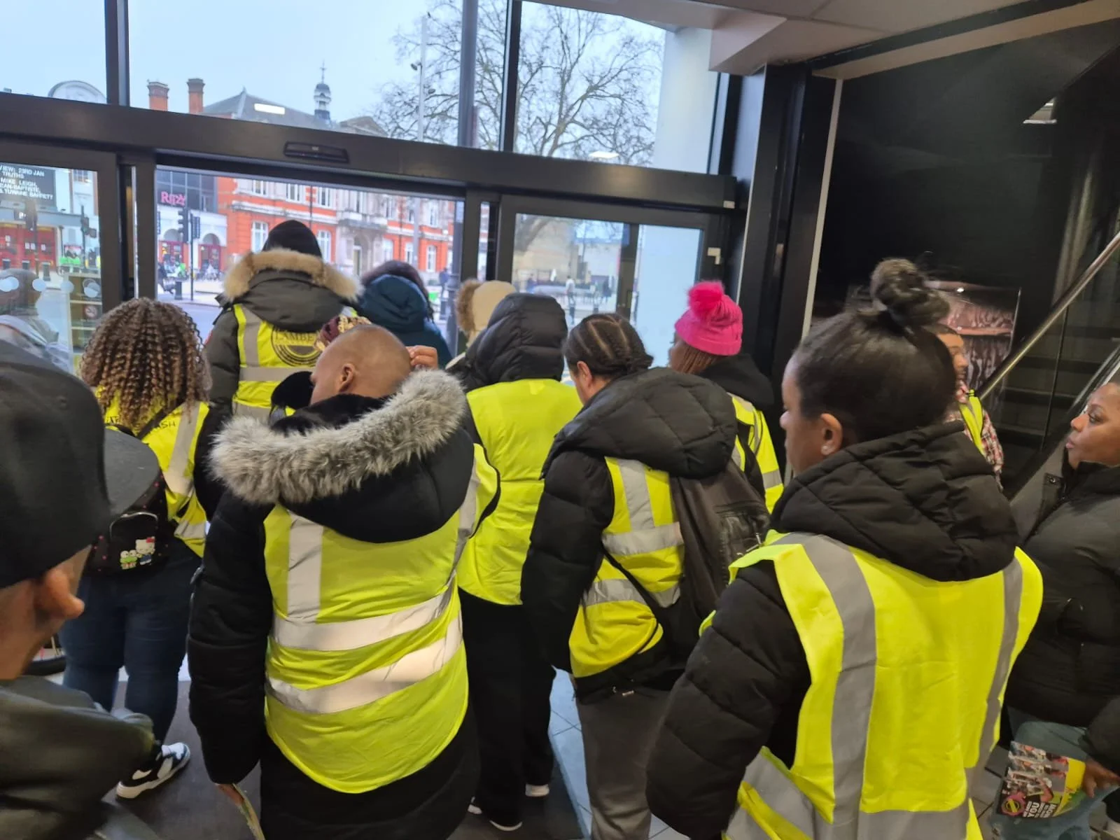 A group of people wearing yellow reflective safety vests standing inside a building near the entrance, some looking outside through glass doors.