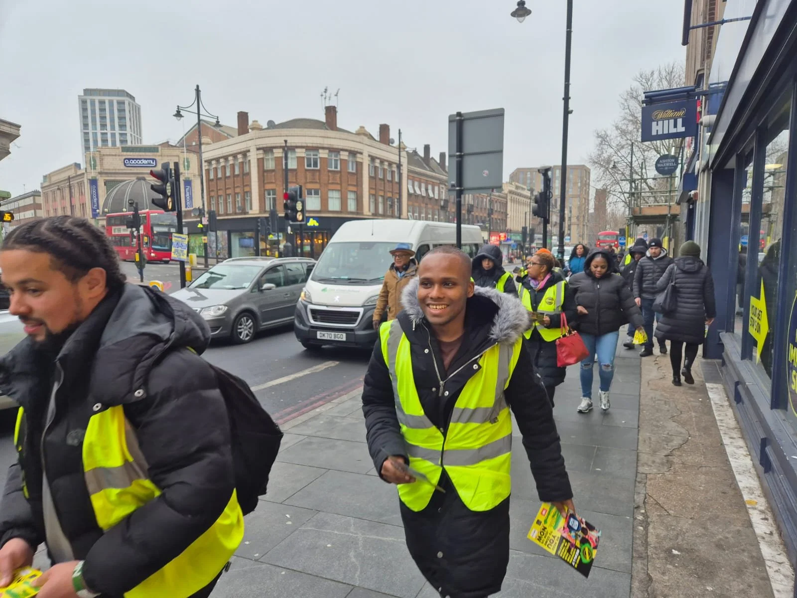 A group of people, many wearing yellow safety vests, walking on a city sidewalk on a cloudy day, with buildings, cars, and a red bus in the background.