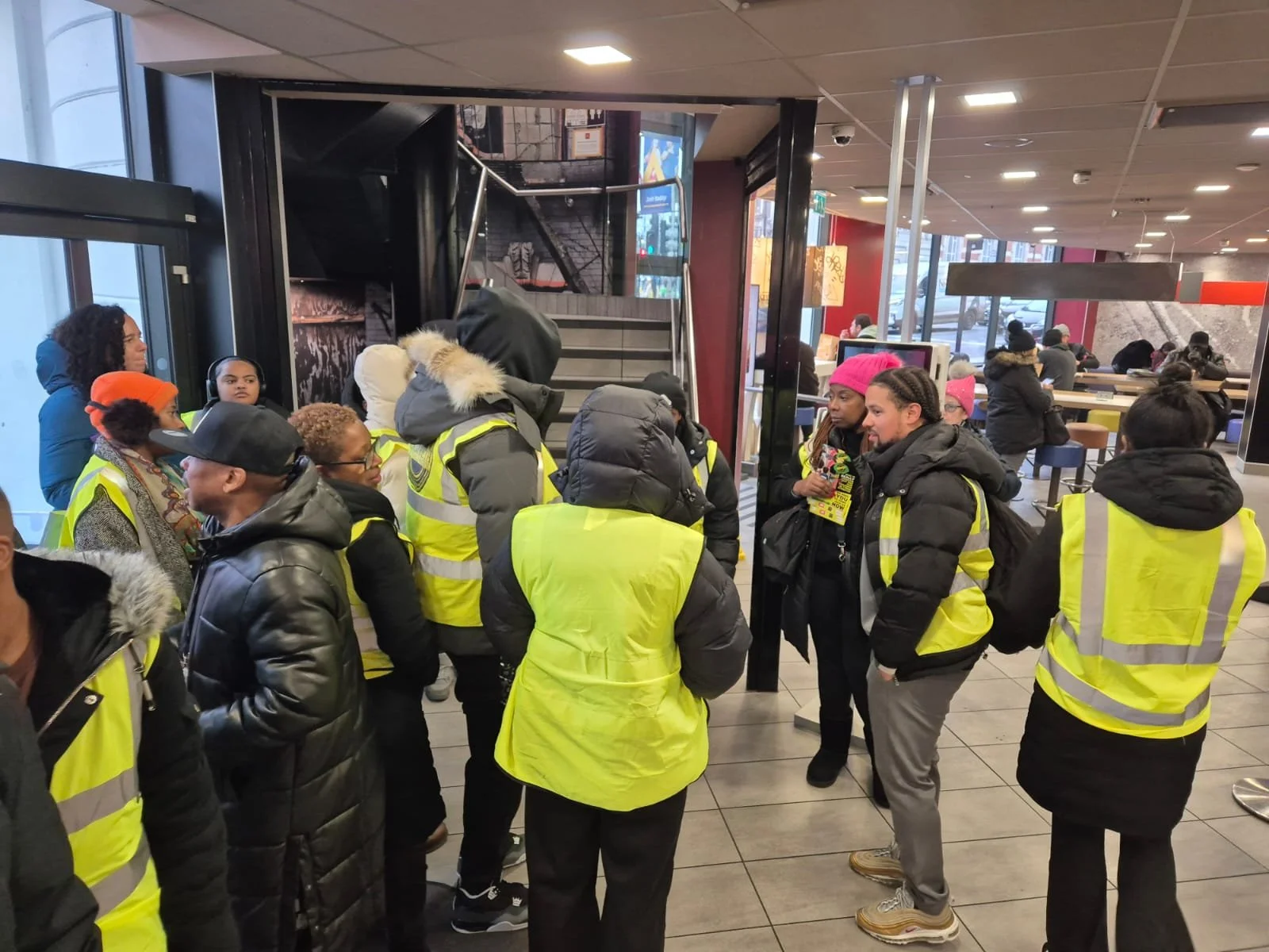 Group of people wearing yellow safety vests gathering inside a building near an entrance, with some holding pamphlets and others using phones, in a busy public space.