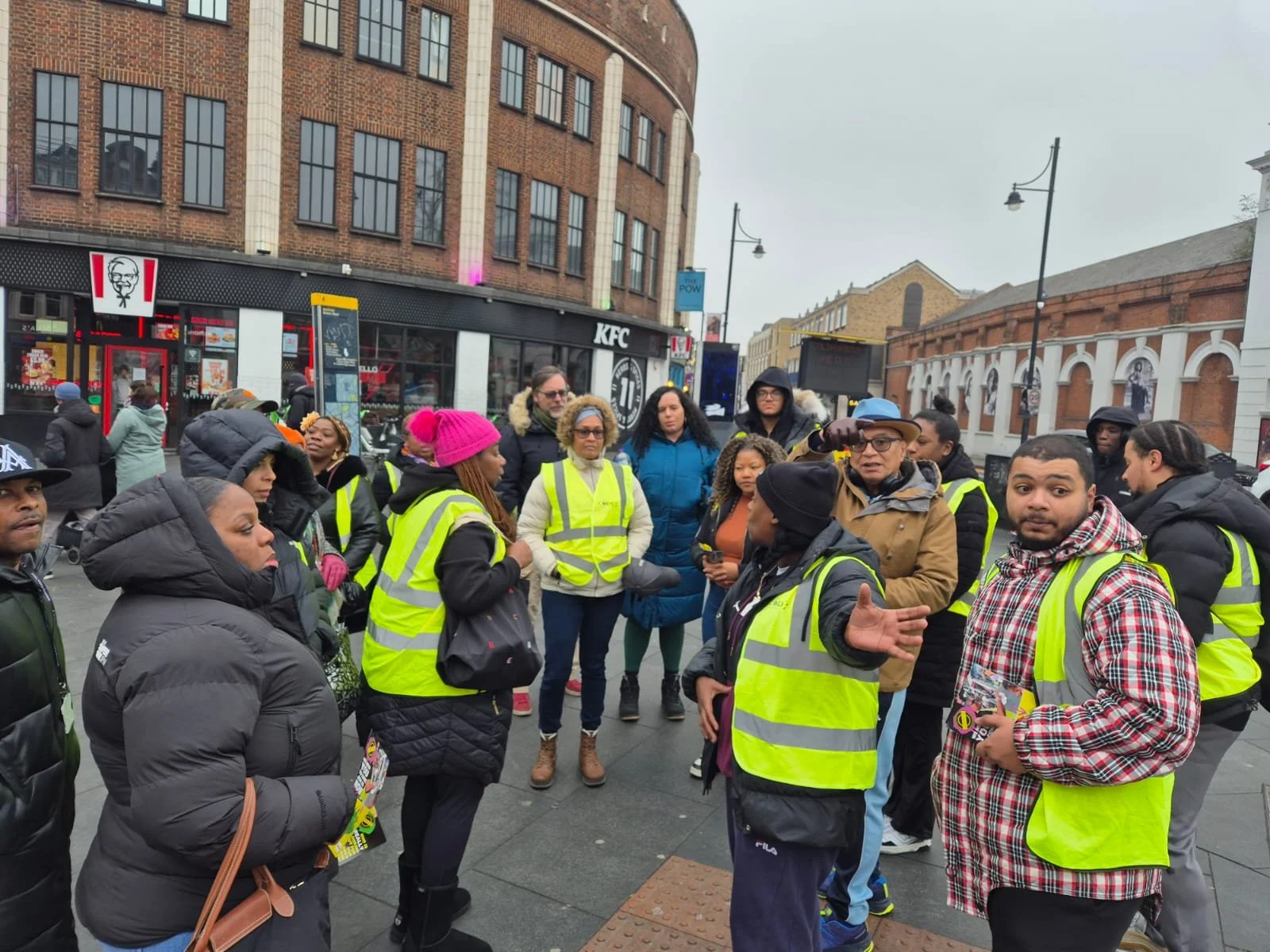 A group of people wearing yellow safety vests gathered outdoors in a city street, listening to a person speaking, with various buildings, including a KFC restaurant, in the background.