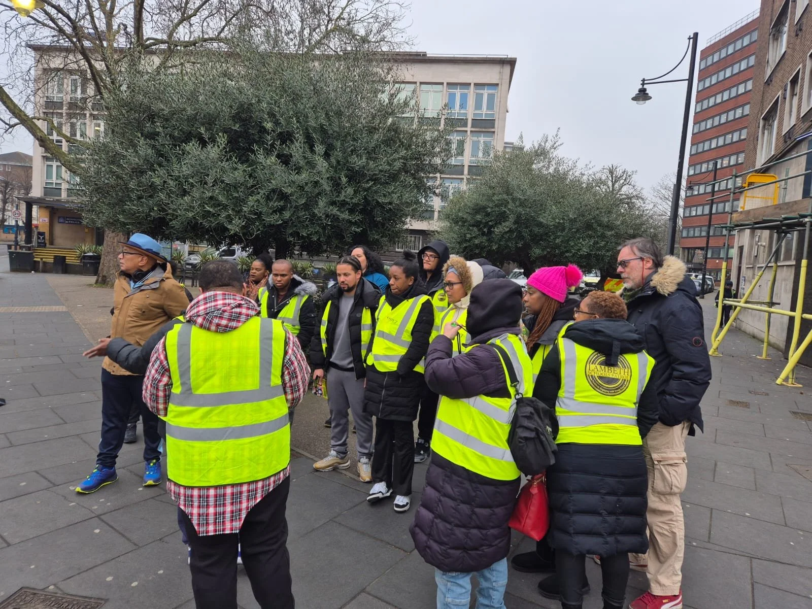 A group of people wearing yellow reflective safety vests gathered outdoors in an urban park or plaza, listening to a person who appears to be leading or instructing them.