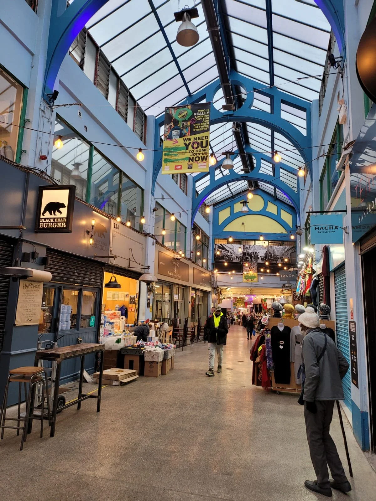 Indoor marketplace with string lights, clothing stalls, and a glass ceiling with blue accents.