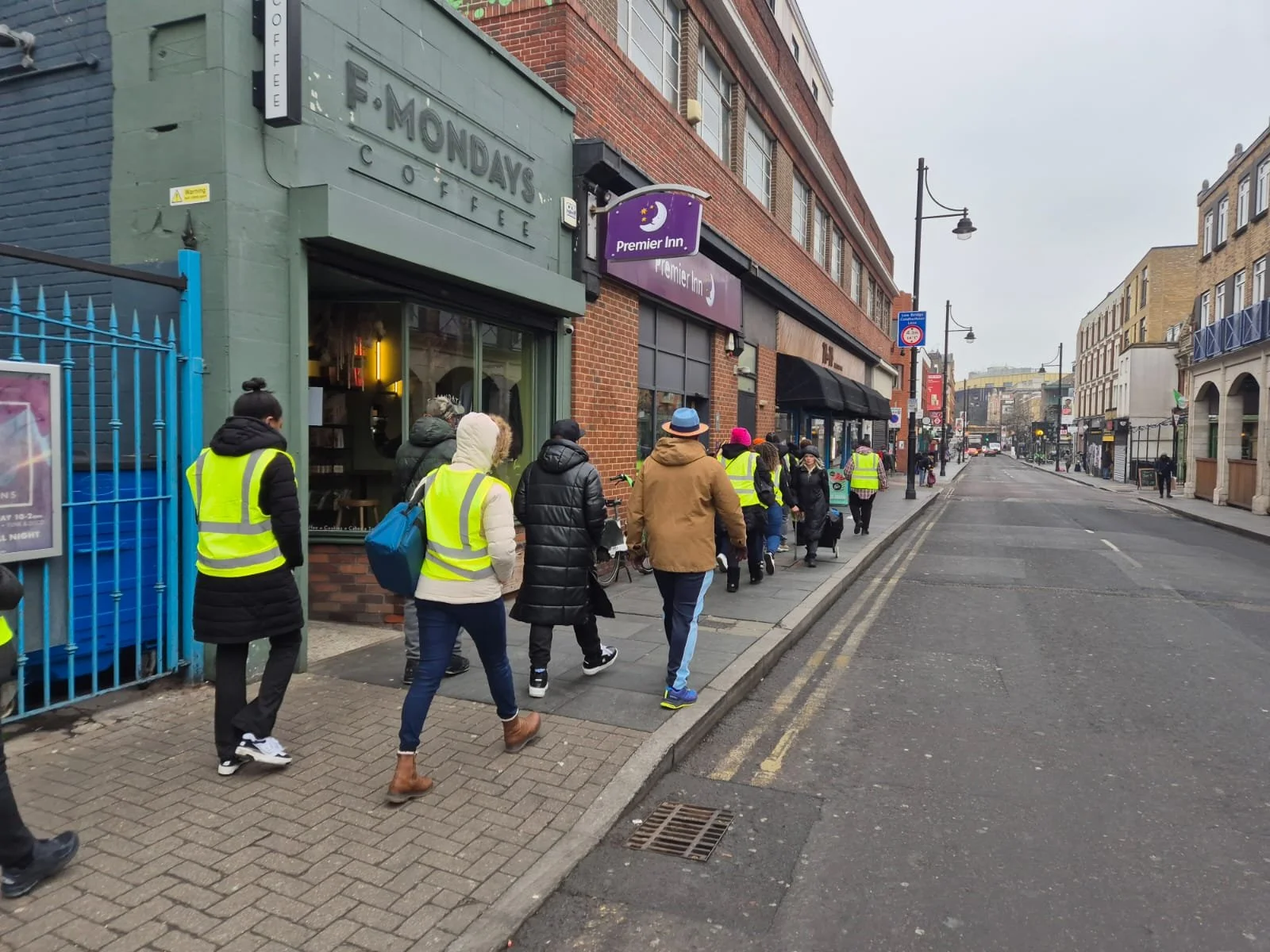 People wearing yellow safety vests walking on a city sidewalk in front of a coffee shop called F-MONDAY'S COFFEE, with a row of buildings along the street, some with black or navy facades and signage for a hotel called Premier Inn.
