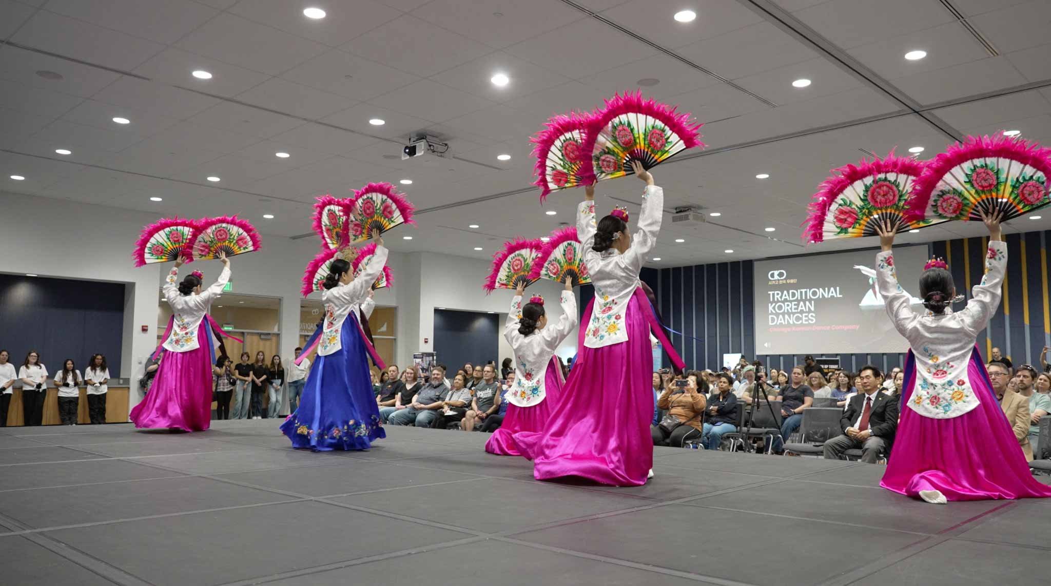 Six women in traditional Korean dresses perform a fan dance on stage, holding colorful fans with pink feathers, in front of an audience at an indoor event. A sign in the background reads "Traditional Korean Dances."